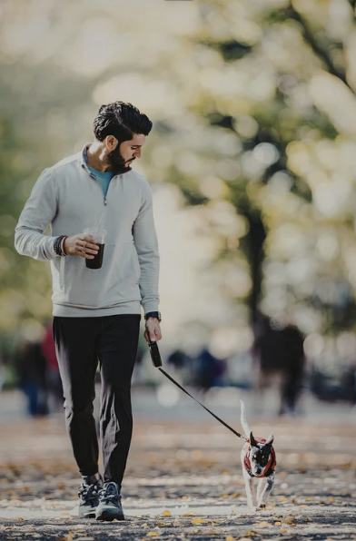 A man walking a small dog on a leash in a park during autumn, holding a beverage.