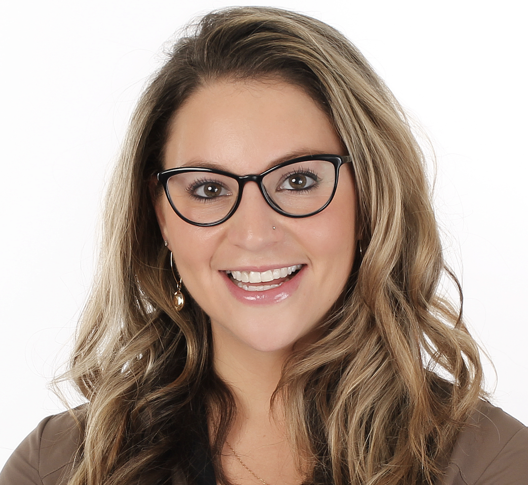 A woman with long wavy hair, wearing glasses, earrings, and a brown top, smiling at the camera with a plain white background.