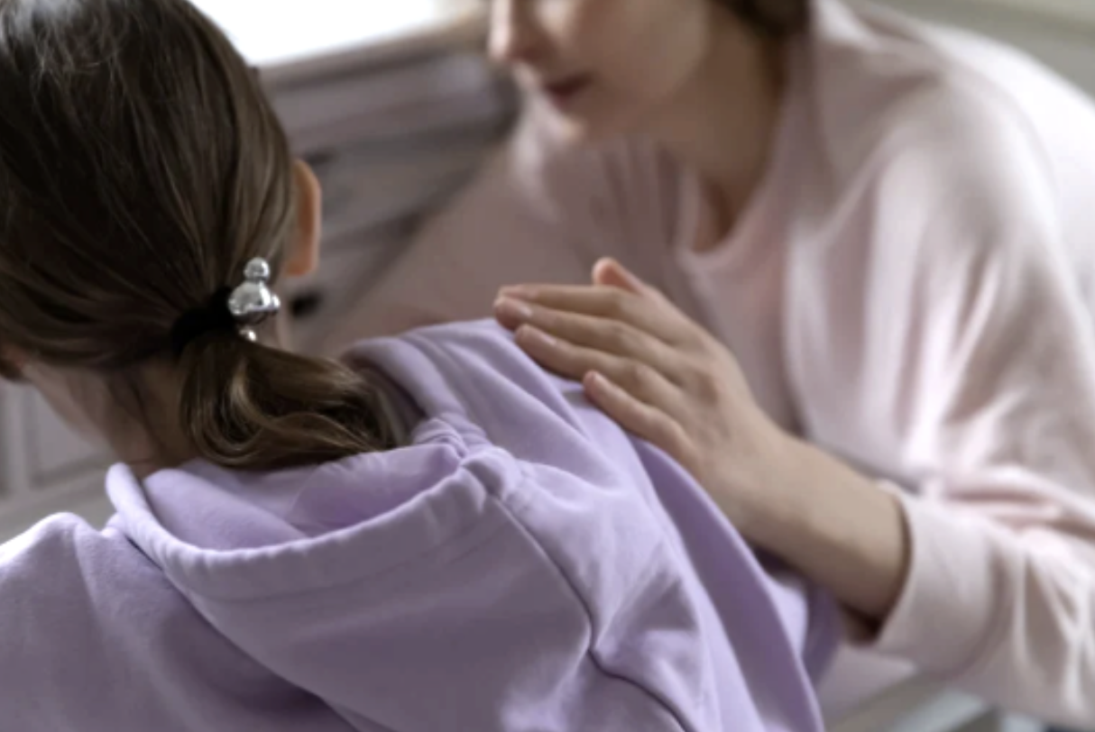 A woman comforting a girl by placing her hand on her shoulder in an indoor setting.
