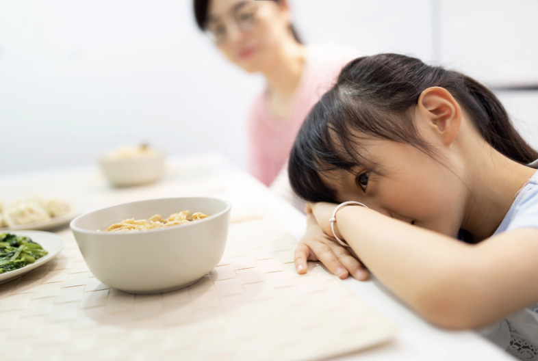 A girl resting her head on a table in front of a bowl of noodles, with a woman blurred in the background, near a table with other dishes.