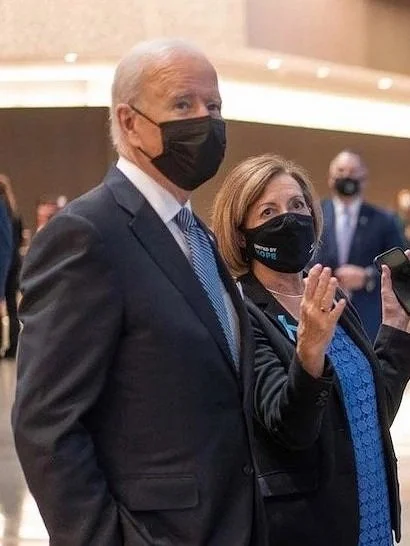 Joe Biden and a woman are standing together at an indoor event, both wearing black face masks. The woman is waving and holding a phone, while Biden is looking to the side.