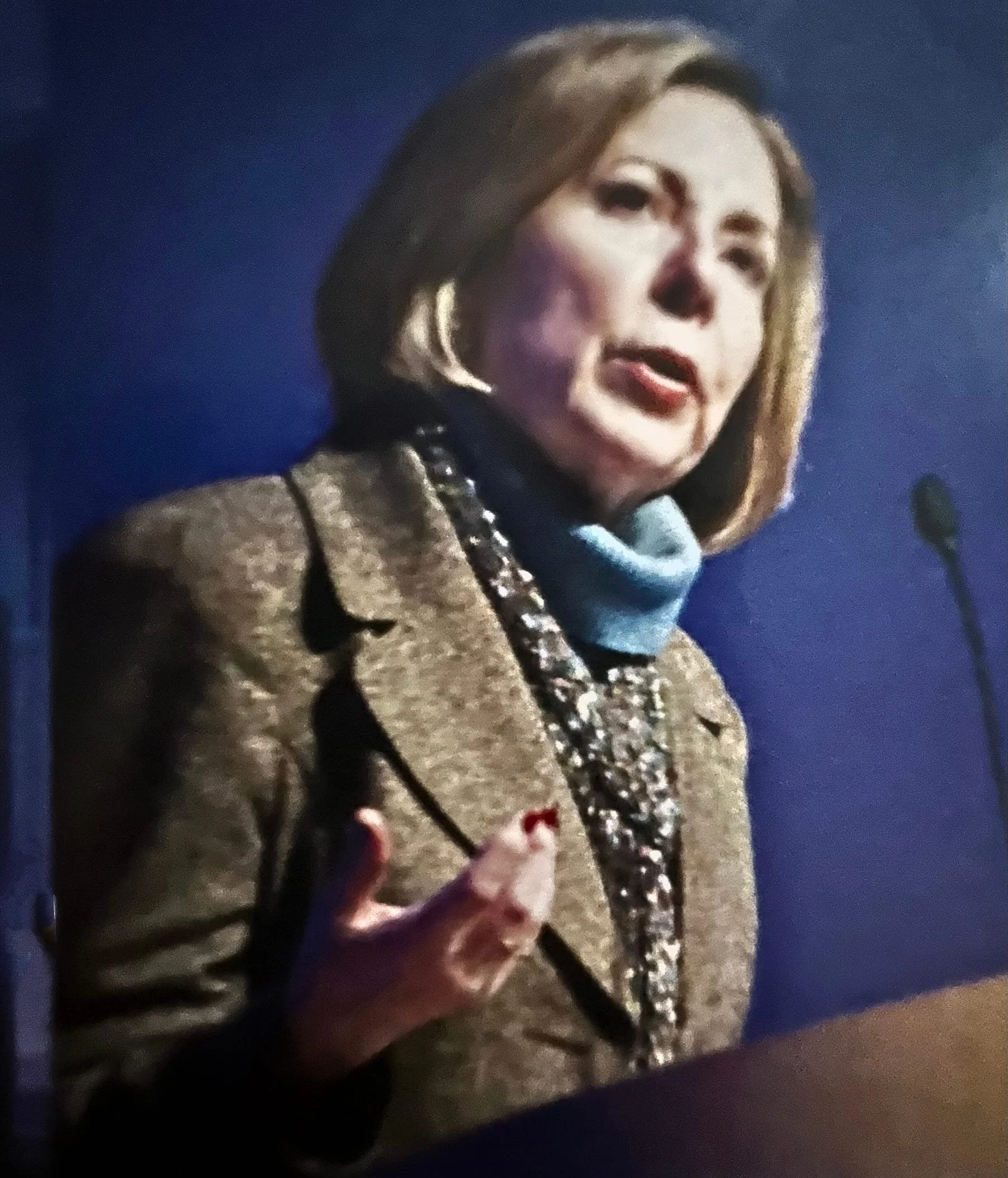 Alice M. Greenwald speaking at a podium with a blue background, dressed in a blazer with a scarf and layered jewelry.