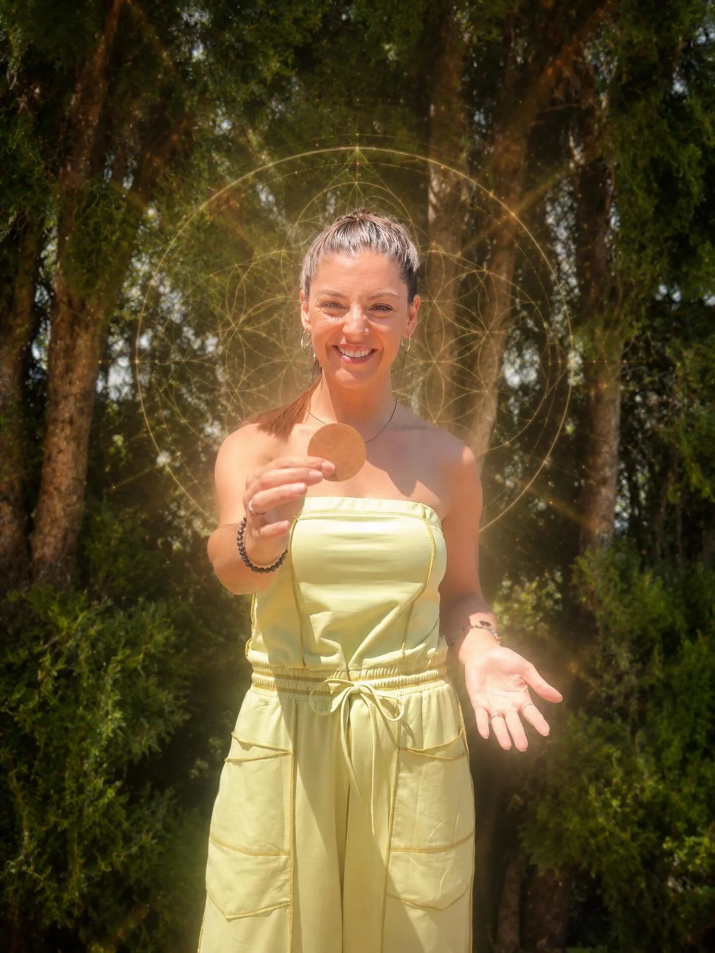 A smiling woman in a yellow strapless jumpsuit standing outdoors with trees in the background. She is holding a small round object in one hand and has her other hand extended open. There is a glowing geometric pattern overlay around her head.