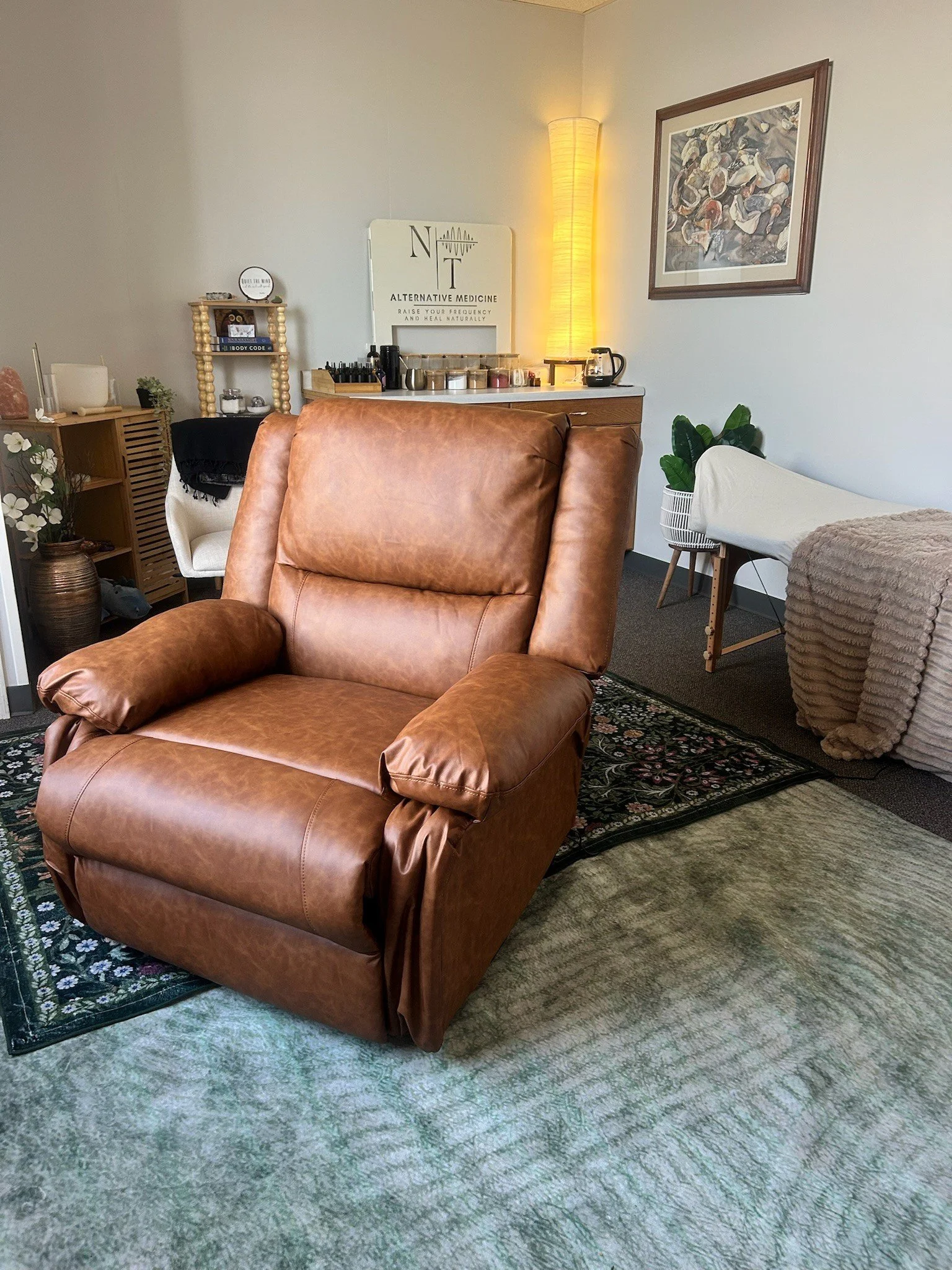 A cozy living room with a brown leather armchair, a brass vase with white flowers, a white chair with a black blanket, a tall yellow floor lamp, a wooden side table, and a wall-mounted framed artwork of seashells. Carpeted floor with a patterned rug.