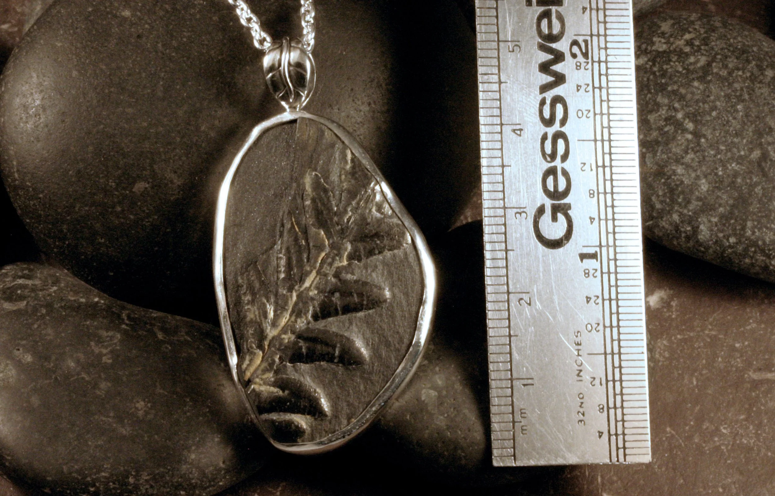 A sterling silver black shale fern fossil pendant next to a ruler.