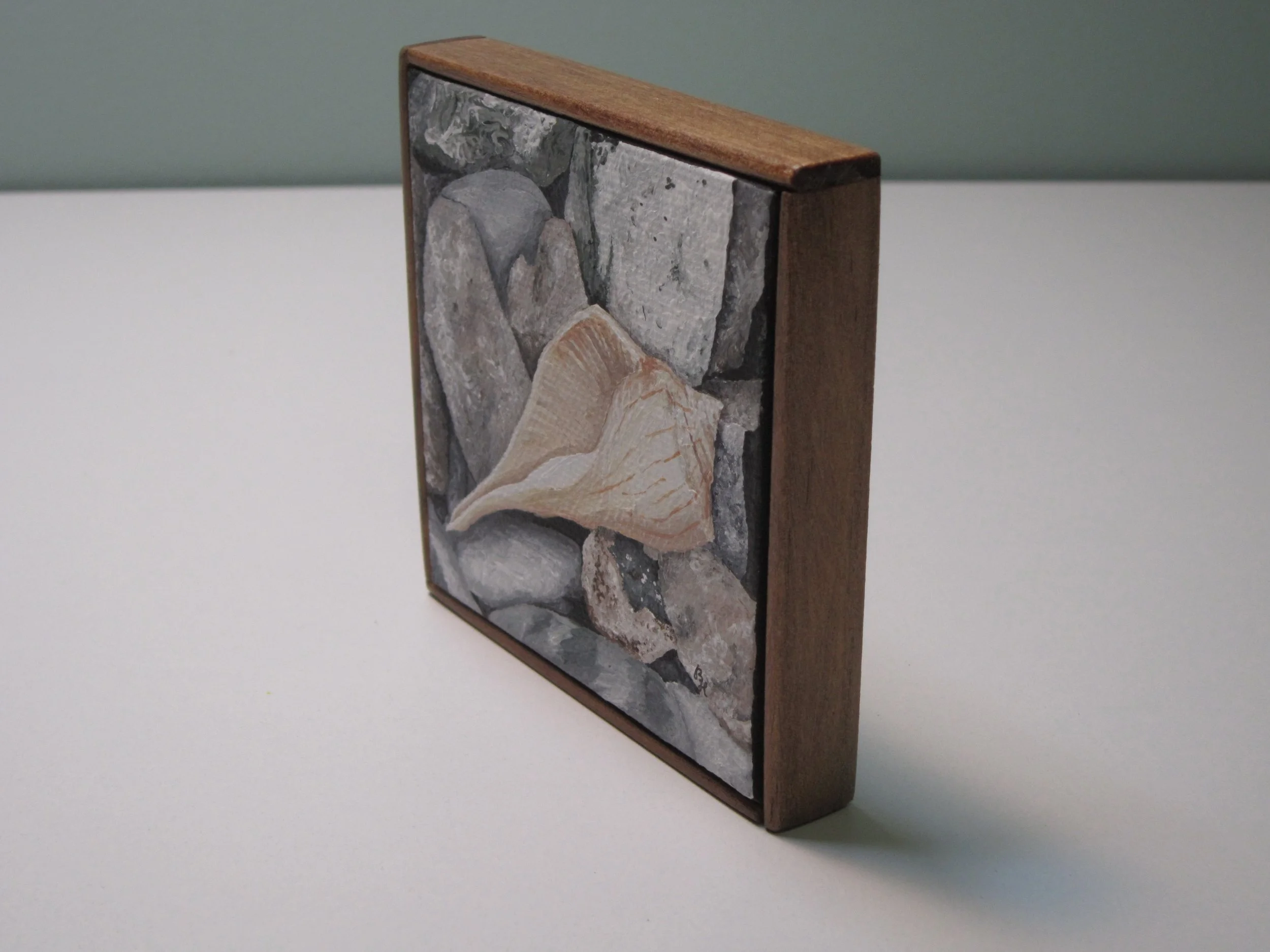 An angled view of a mini acrylic painting of a seashell on rocks.
