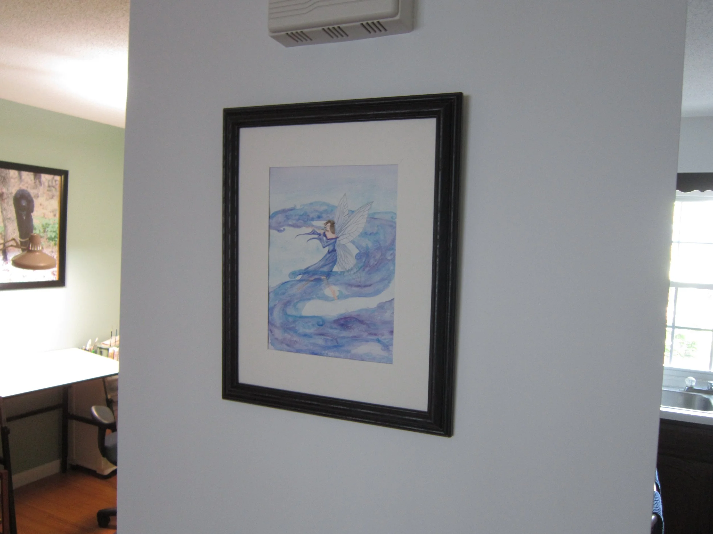 Hanging on a wall is a framed watercolor painting of a fairy blowing out dream clouds that are swirling around her.
