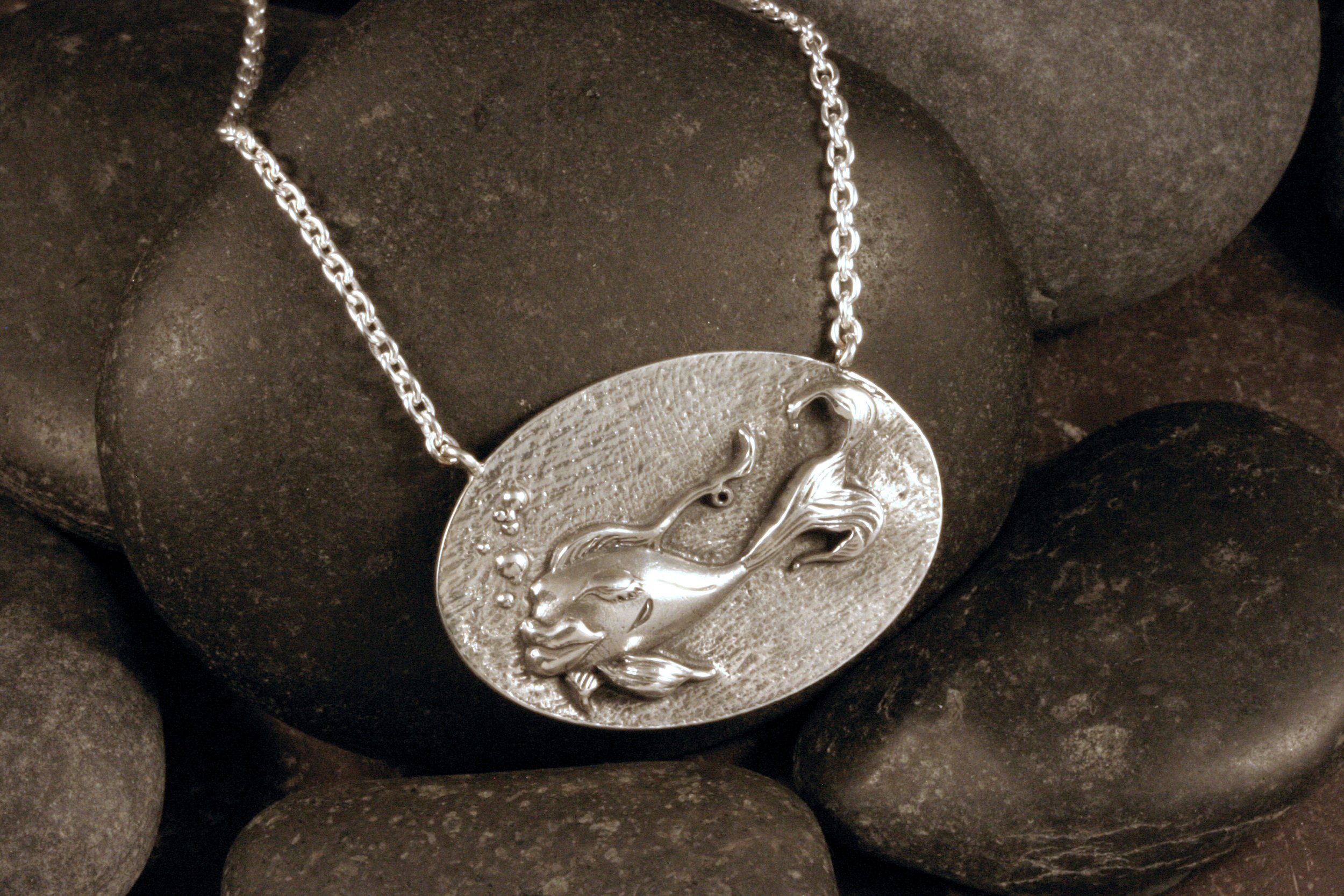 A sterling silver necklace with an oval featuring a relief sculpture of a stylized fish with big lips is draped over a pile of black rocks.