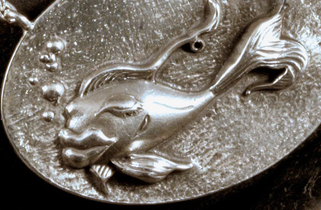 A detail close up image of a sterling silver fish relief sculpture that has big lips and is posed on a textured oval on a necklace.