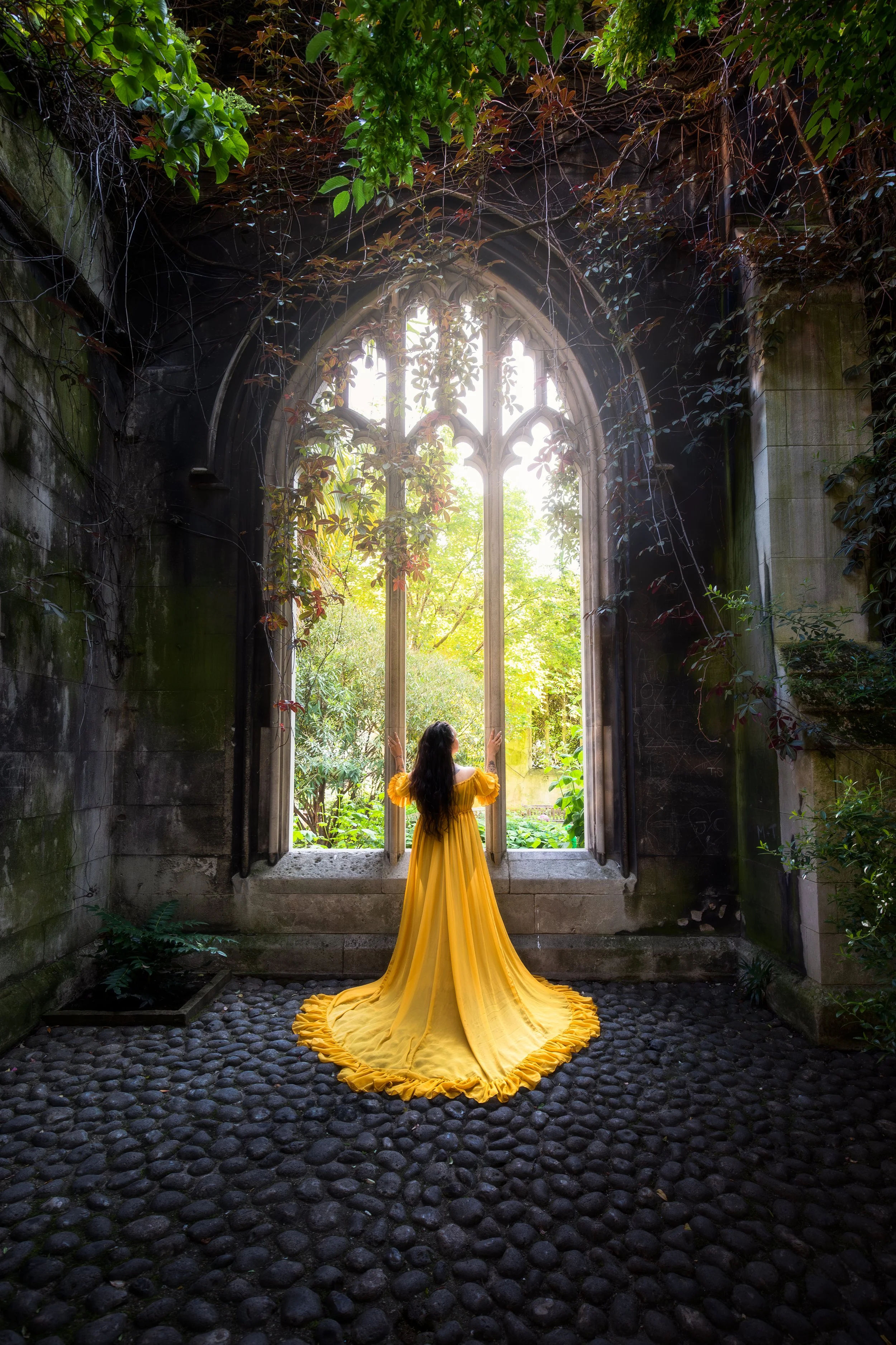A woman in a long yellow dress standing inside a dark, ivy-covered stone structure with a large Gothic-style arched window, looking out to a lush green garden.