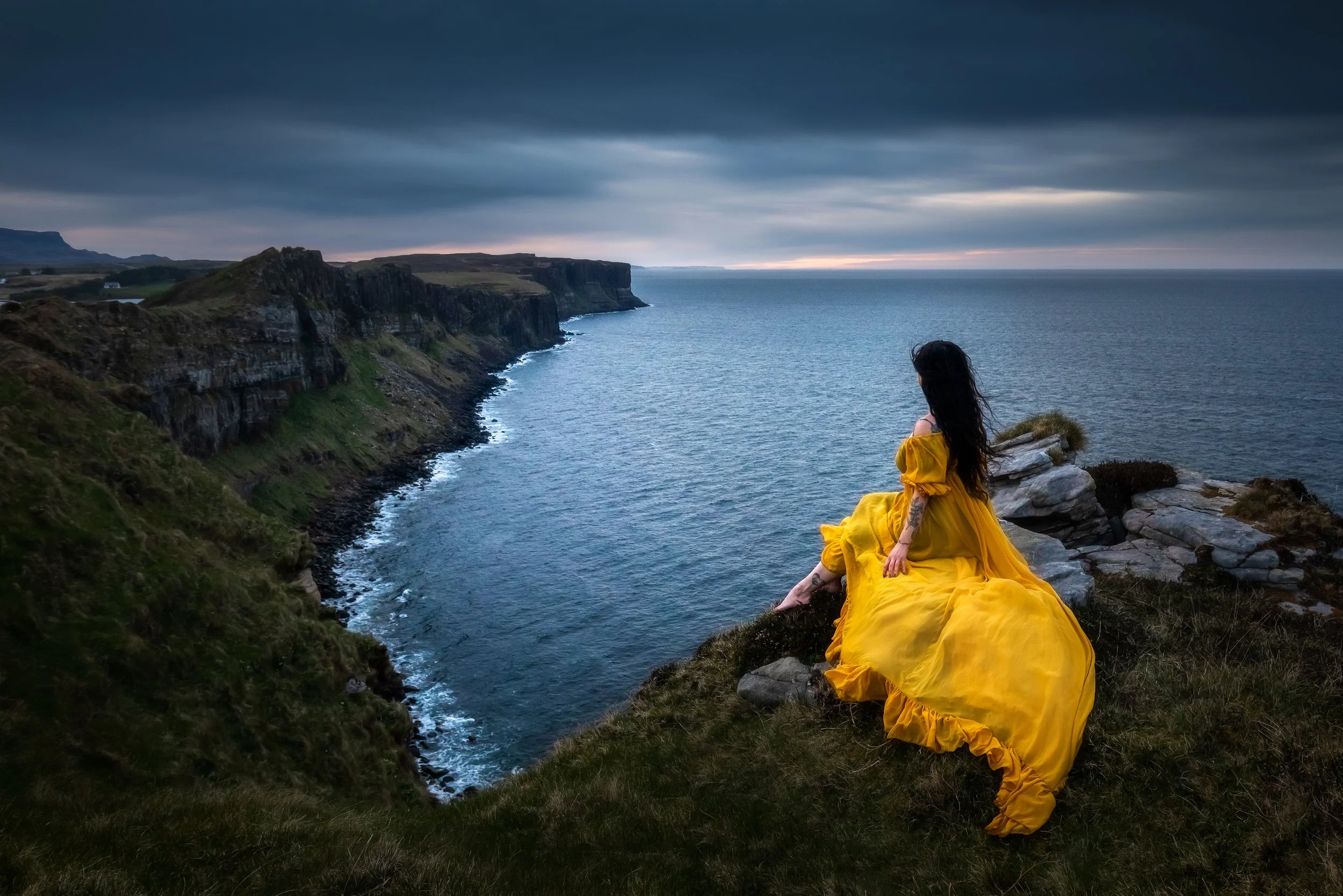 A woman in a flowing yellow dress sitting on rocky grass overlooking a coastal cliff and ocean at dusk, with dark clouds overhead.