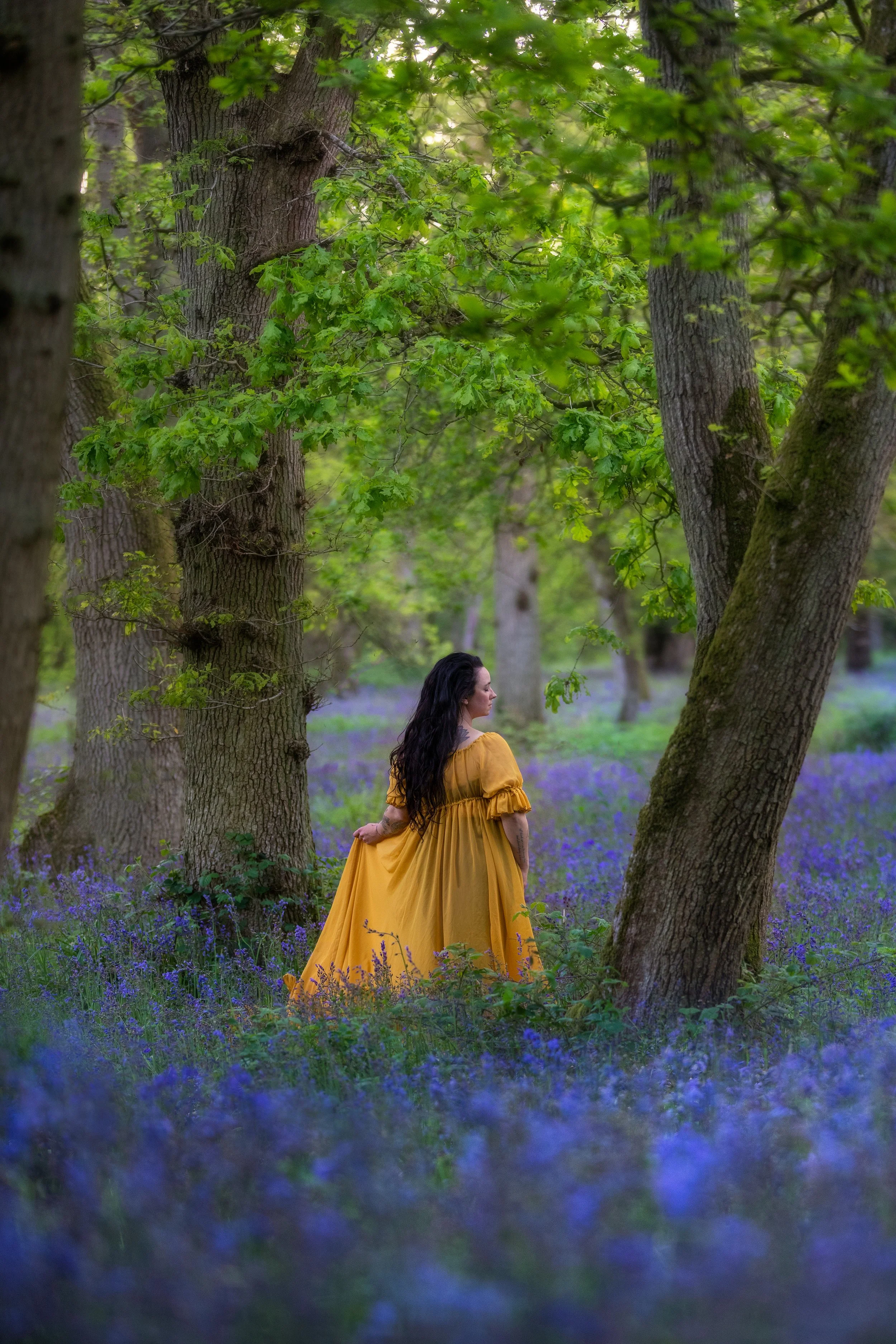 A woman with long dark hair wearing a flowing yellow dress standing in a purple flowered forest, surrounded by tall trees with green leaves.