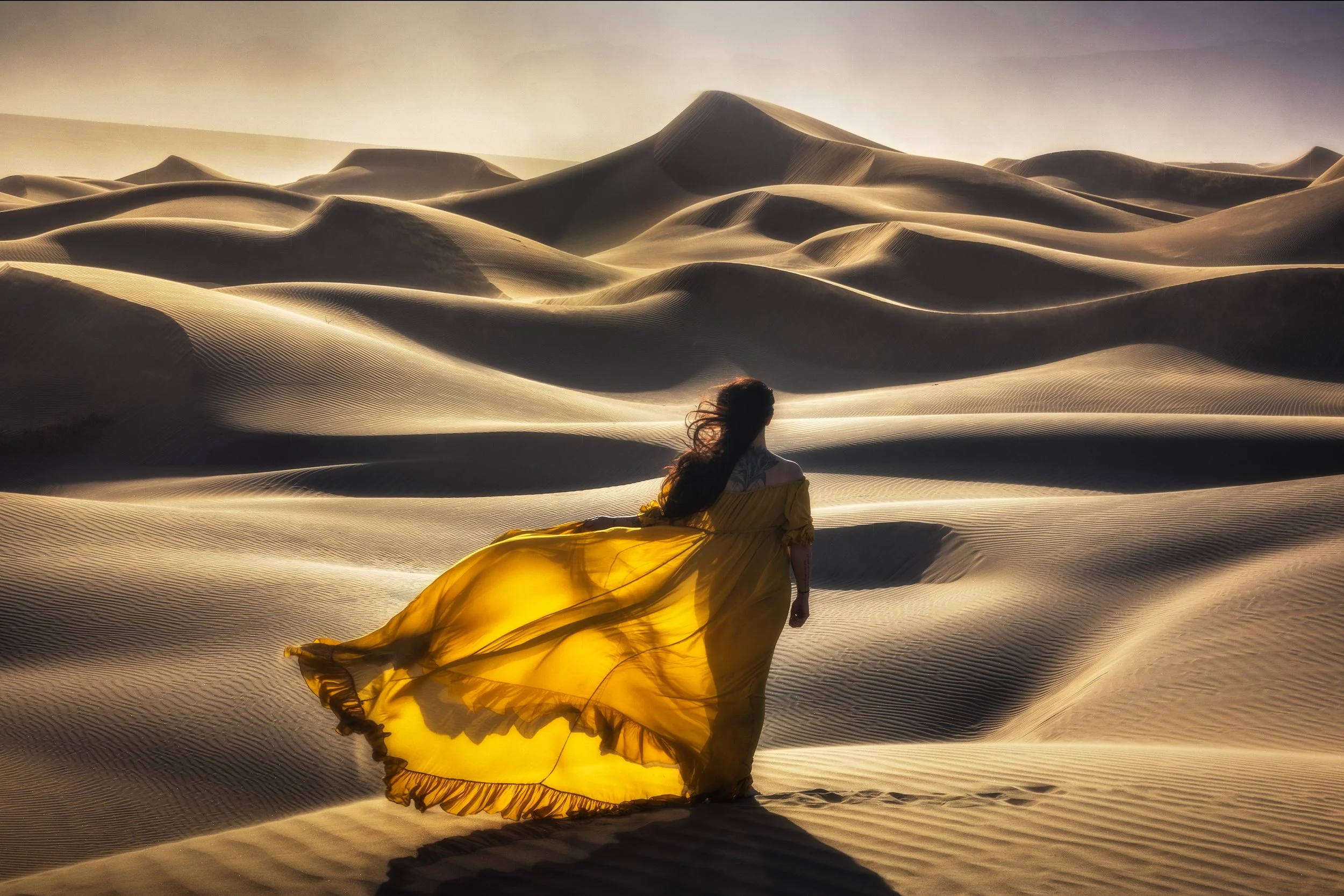 Woman in a flowing yellow dress walking through sand dunes in a desert, with wind blowing her hair.