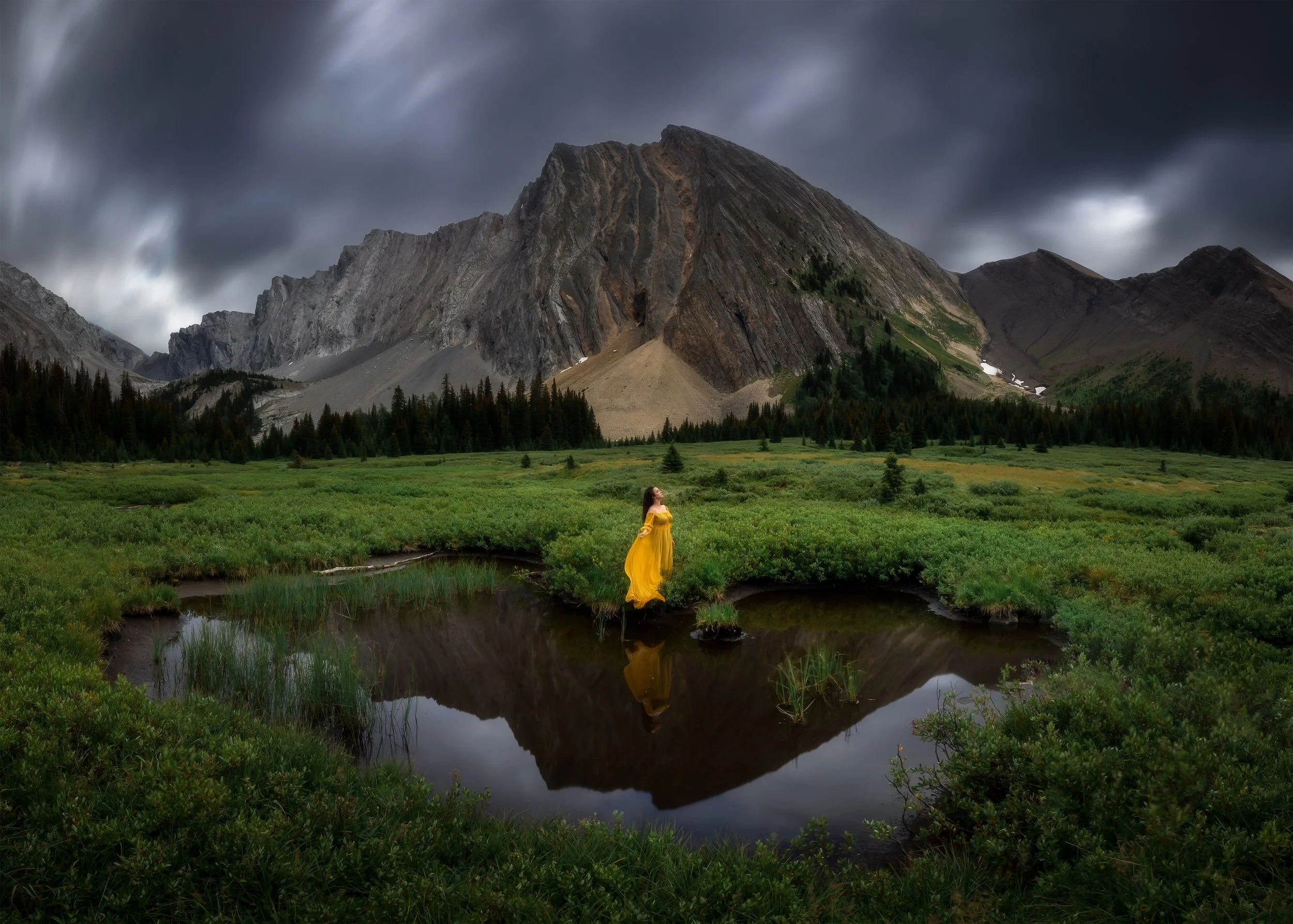 A woman in a yellow dress standing by a reflective pond in a lush green meadow, with a mountain range and dark cloudy sky in the background.