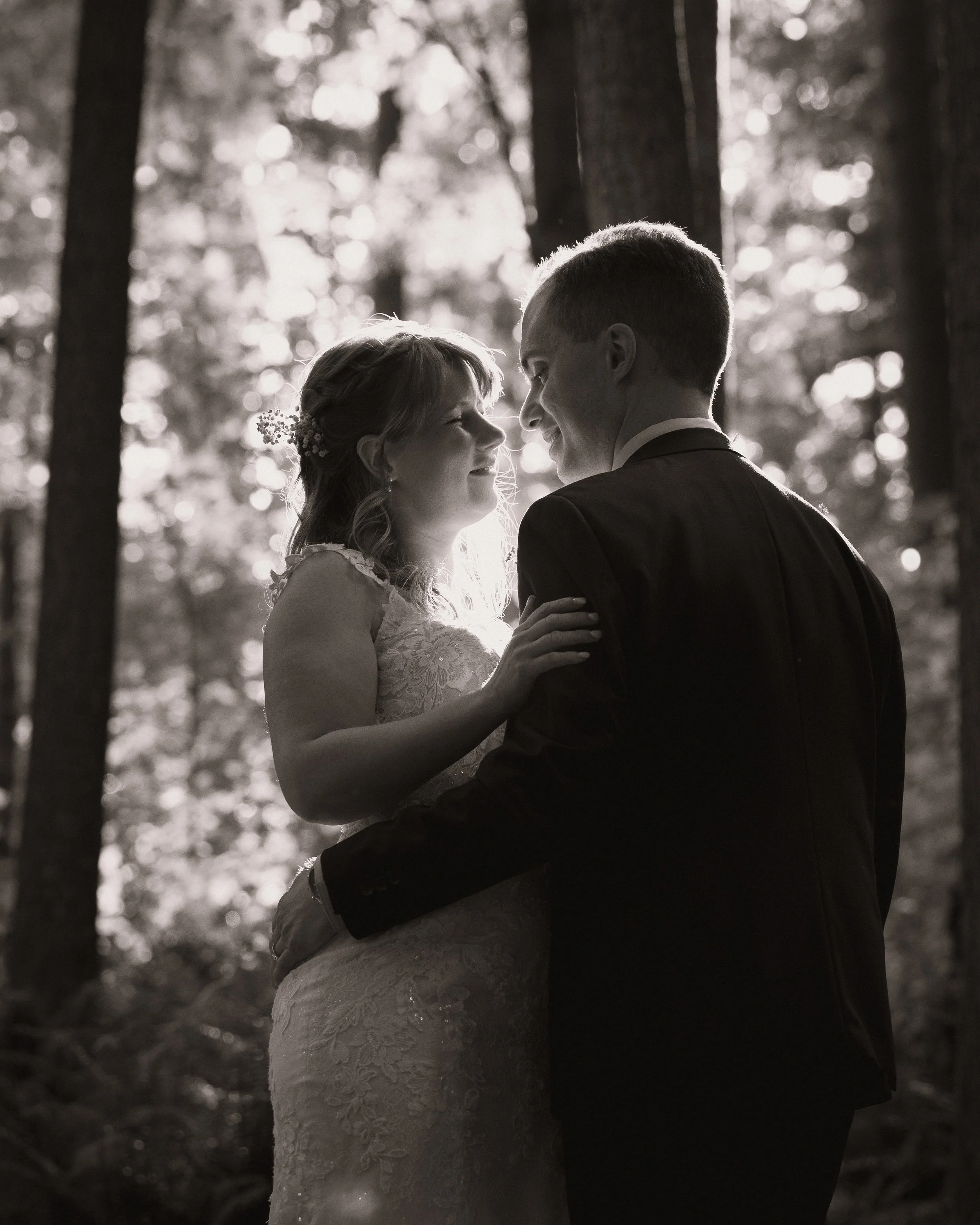 A black and white photo of a bride and groom in a forest at their wedding in Coquitlam, standing close and smiling at each other.