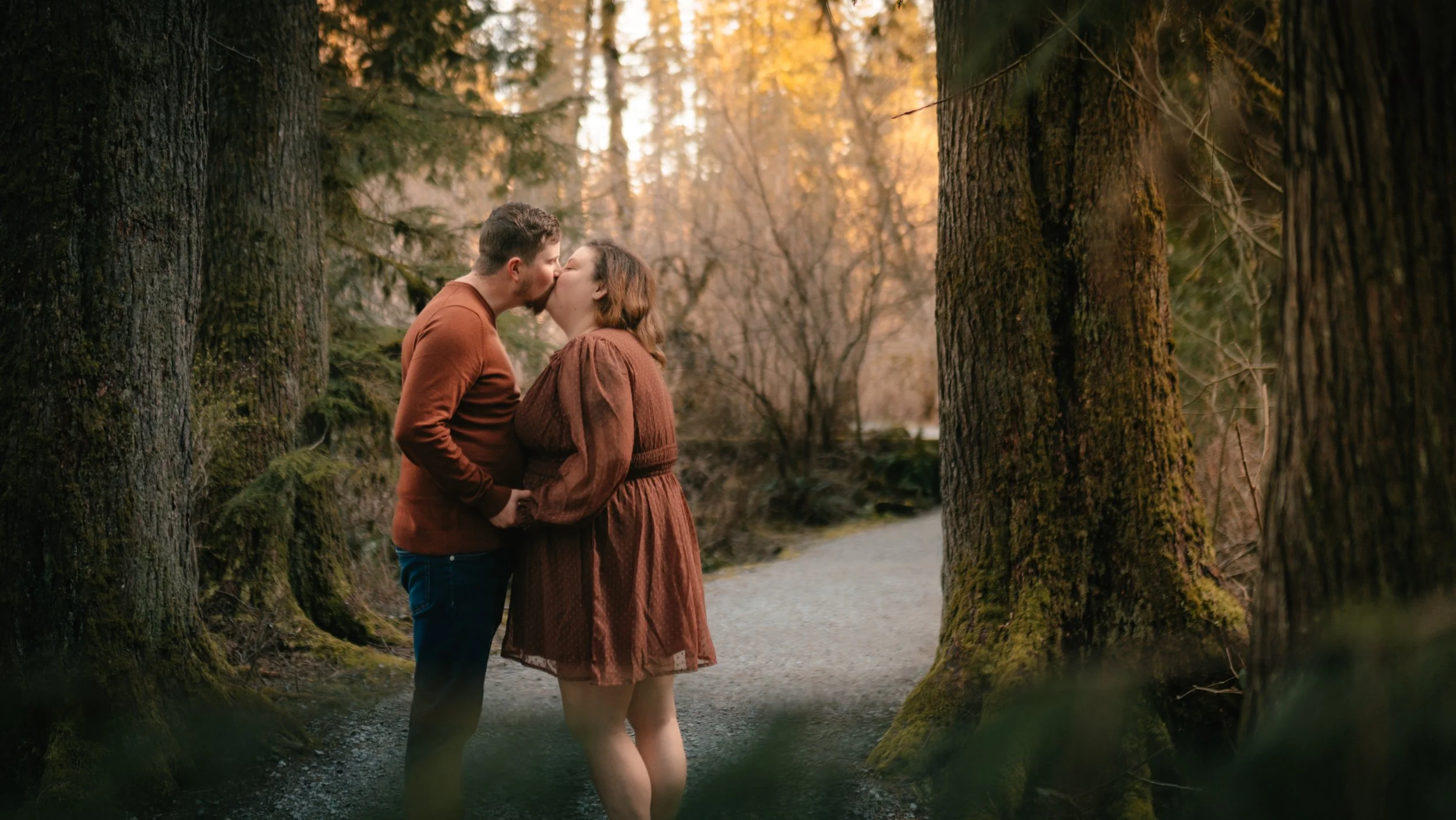 A couple sharing a kiss in a forest during autumn with tall trees, fallen leaves, and a calm stream.