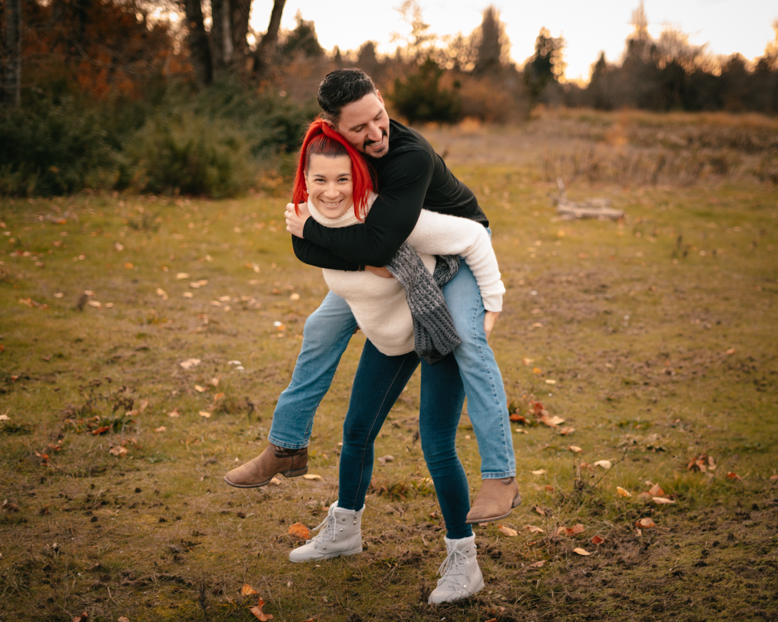 A man giving a piggyback ride to a woman in an outdoor park during autumn, with fall foliage and trees in the background.