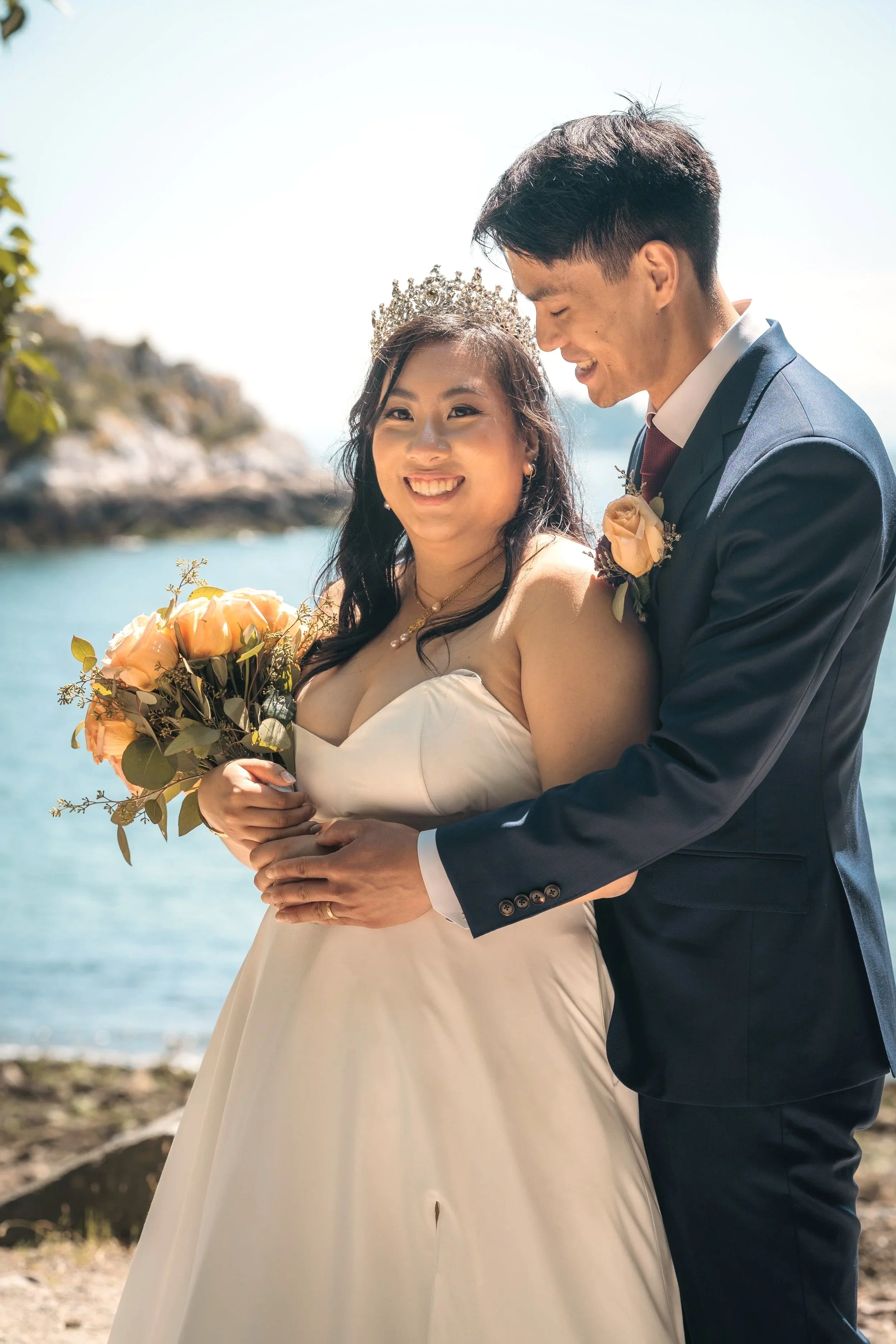 A joyful bride and groom sharing a moment outdoors beside a body of water at their wedding in North Vancouver, with the bride wearing a tiara and holding a bouquet, and the groom in a suit with a boutonniere, under bright sunlight.