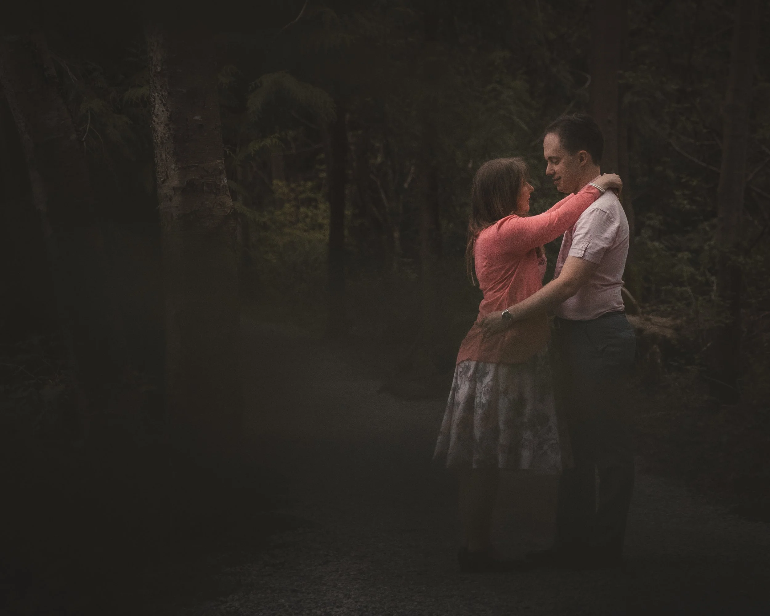 A man and woman are embracing in a forest during nighttime, standing close and looking into each other's eyes.