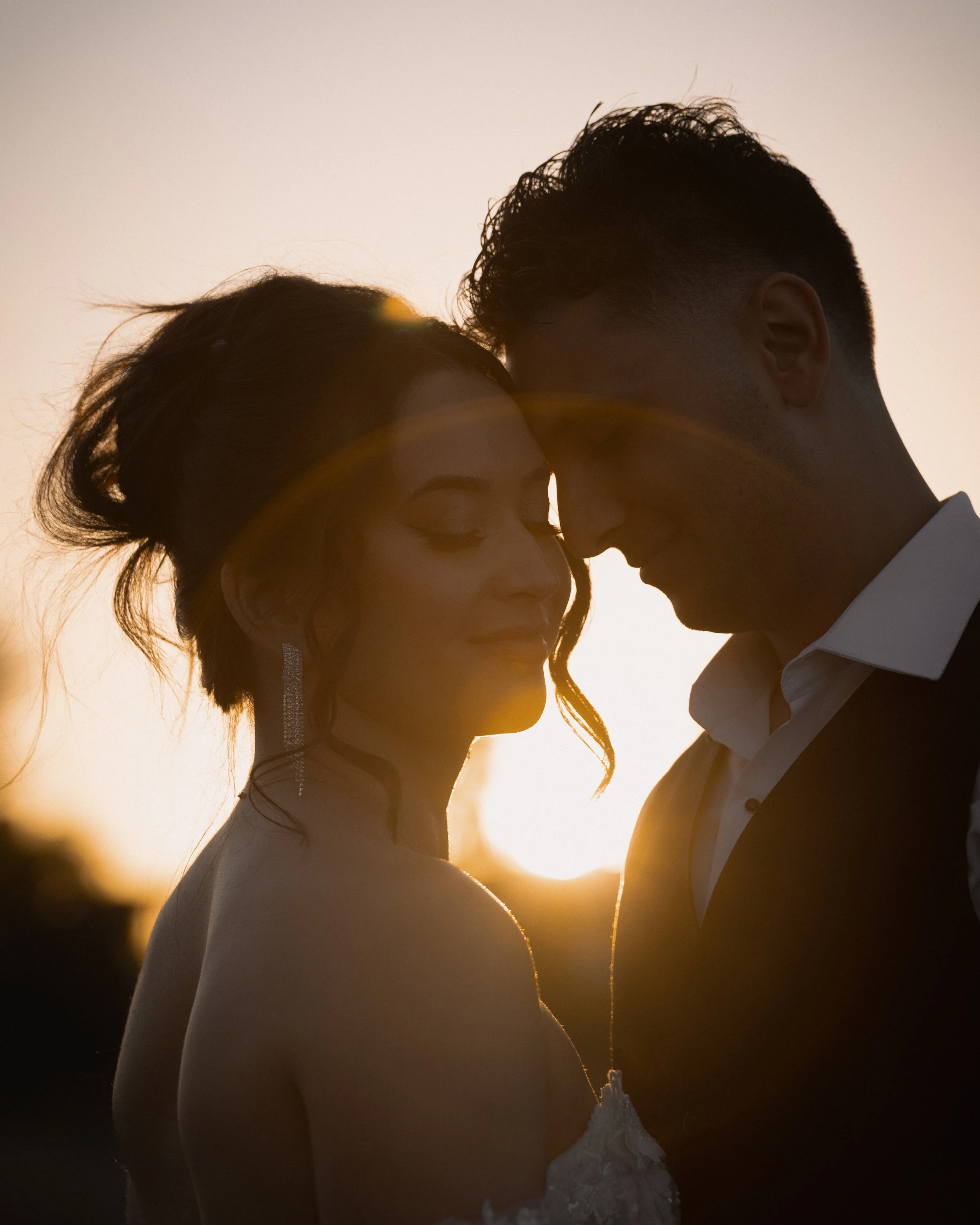 A close-up silhouette of a couple with their foreheads touching during a wedding sunset photo, with a lens flare and a soft sky background.