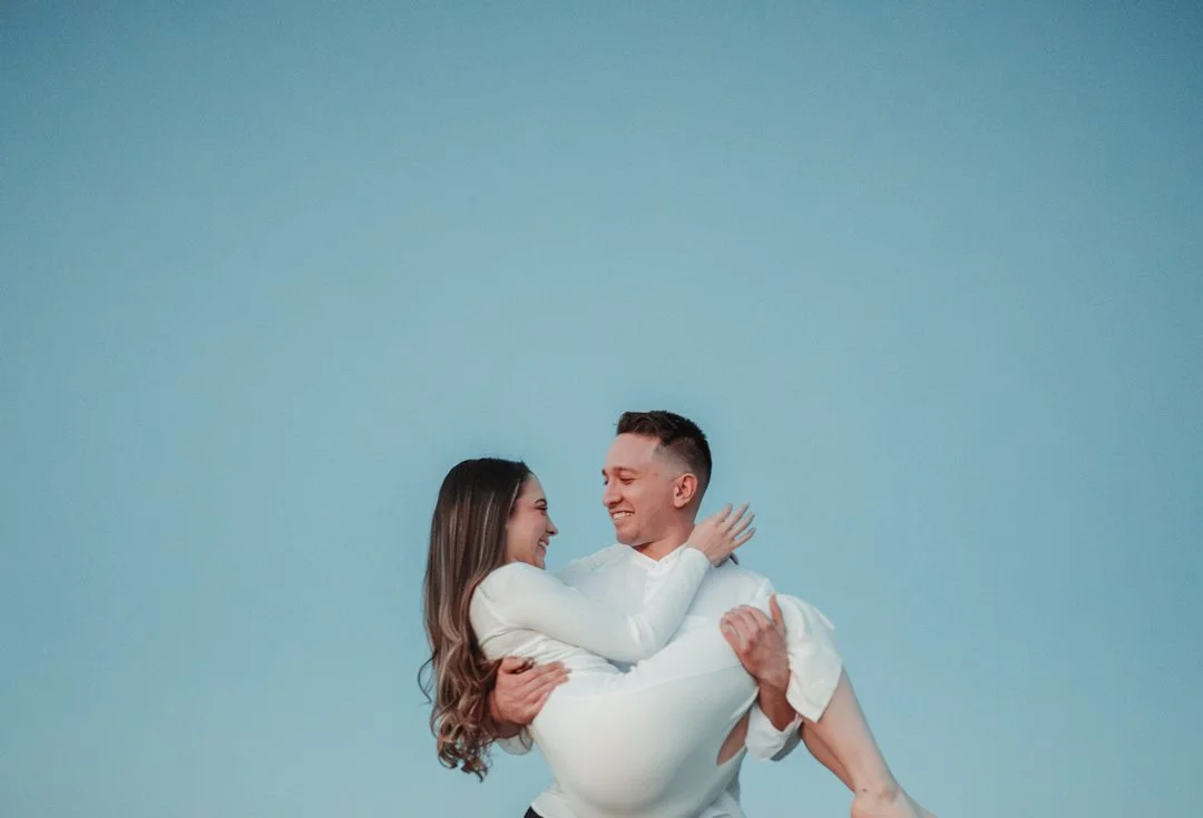 A man holding a woman in his arms against a clear blue sky, both smiling.