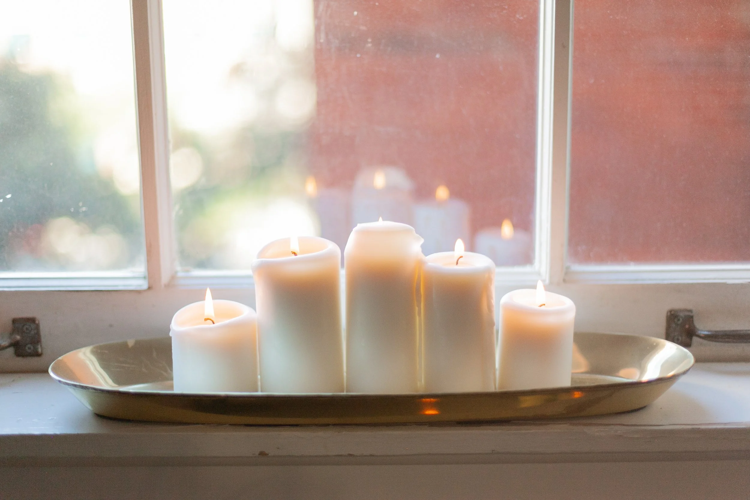 Five white candles on a silver tray placed on a windowsill, with a frosted window and outdoor greenery visible in the background.