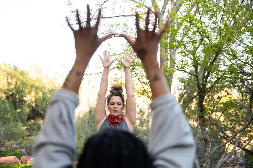 Two women practicing Kundalini yoga outdoors among trees, with one woman in a gray top and a red bandana guiding the other woman in a black top with her hands raised above her head.
