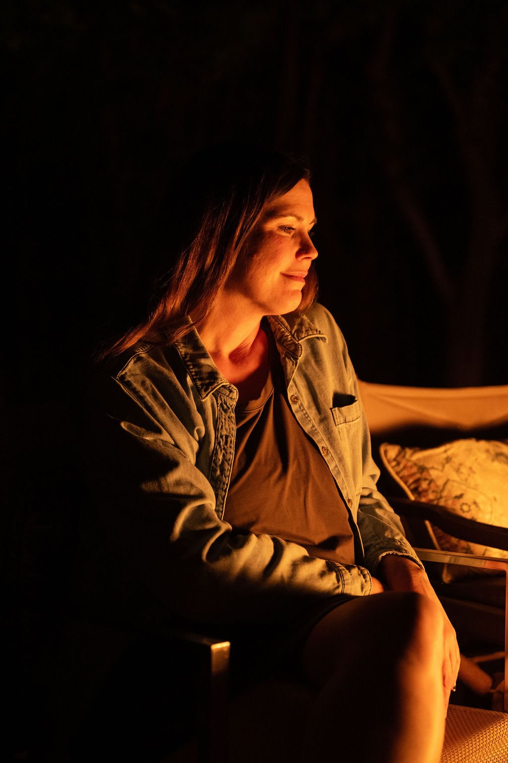 A woman sitting by a campfire at night, smiling softly, illuminated by the warm glow of the fire.