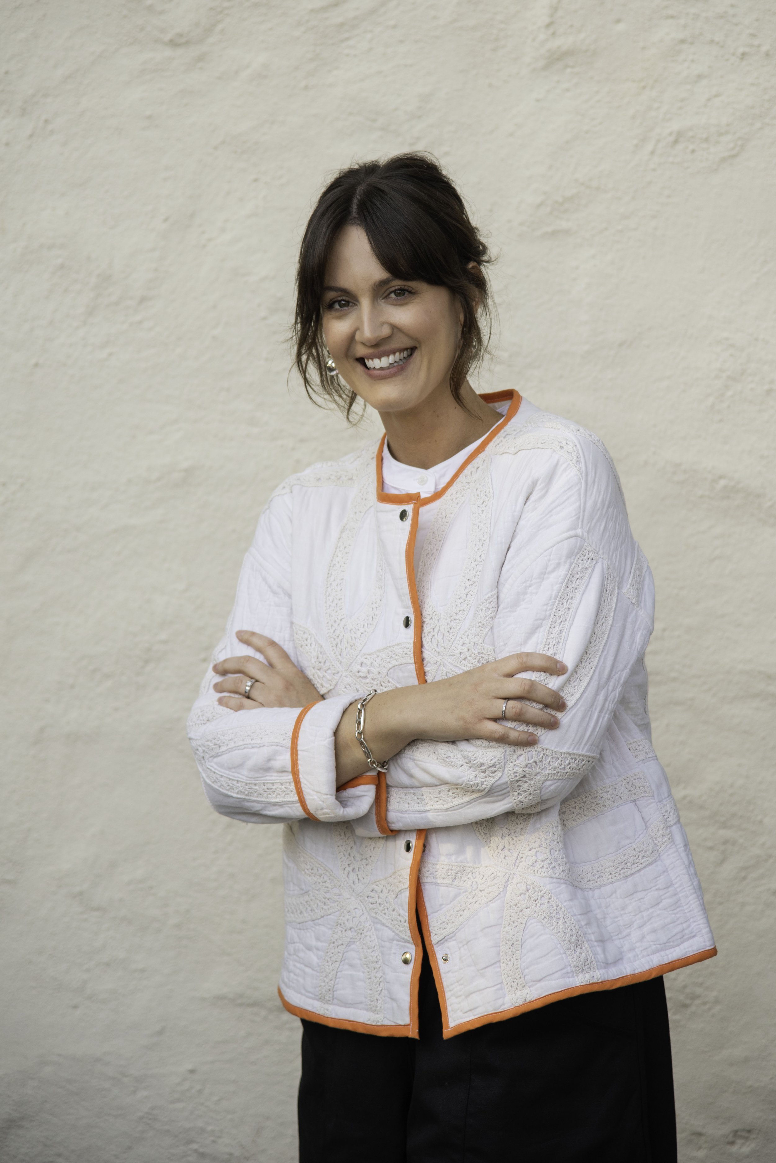 A woman with short brunette hair, smiling, wearing a white jacket with orange piping, and crossing her arms in front of a beige textured wall.