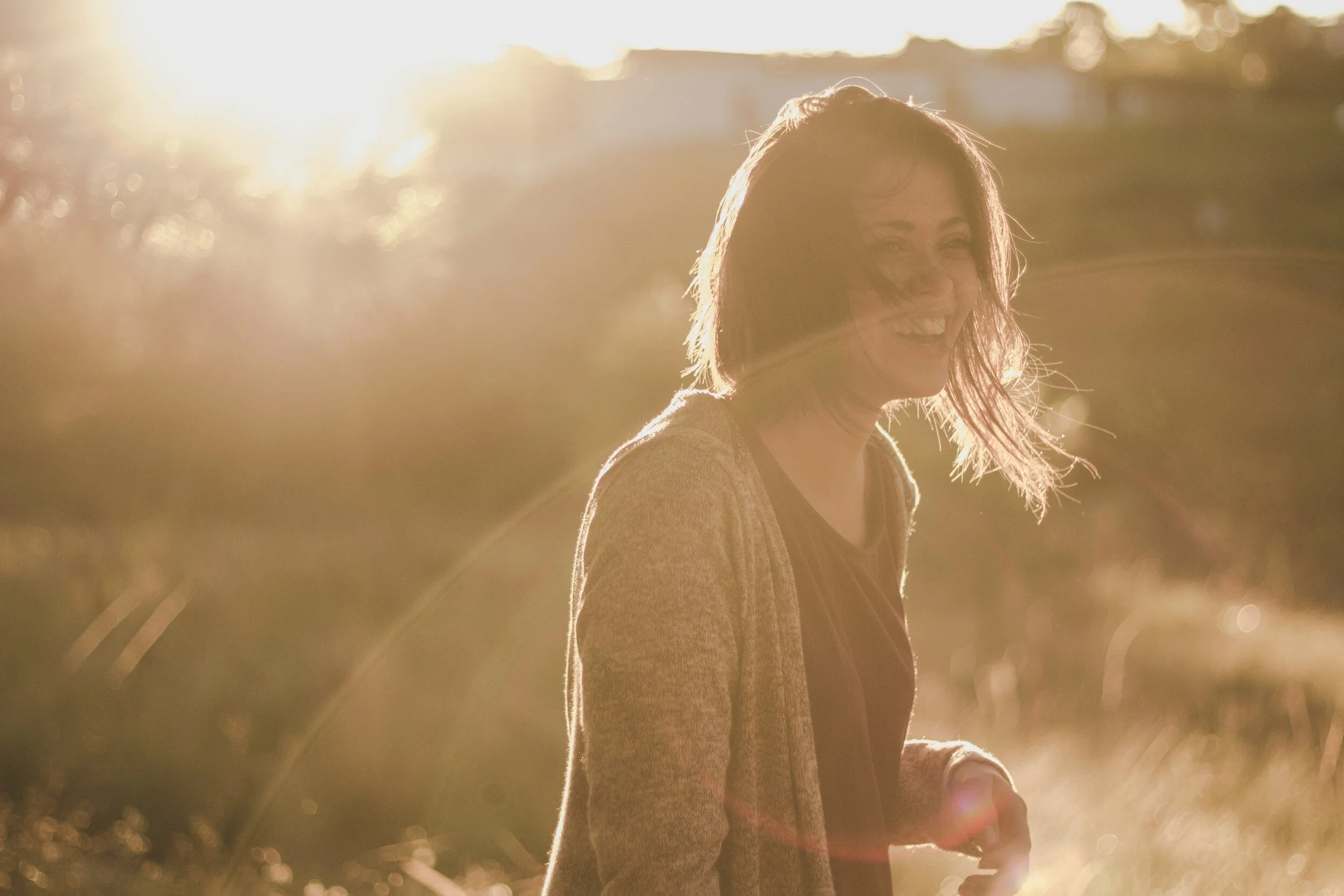 Smiling person backlit by sunset in grassy field, wearing cardigan, with windblown hair.