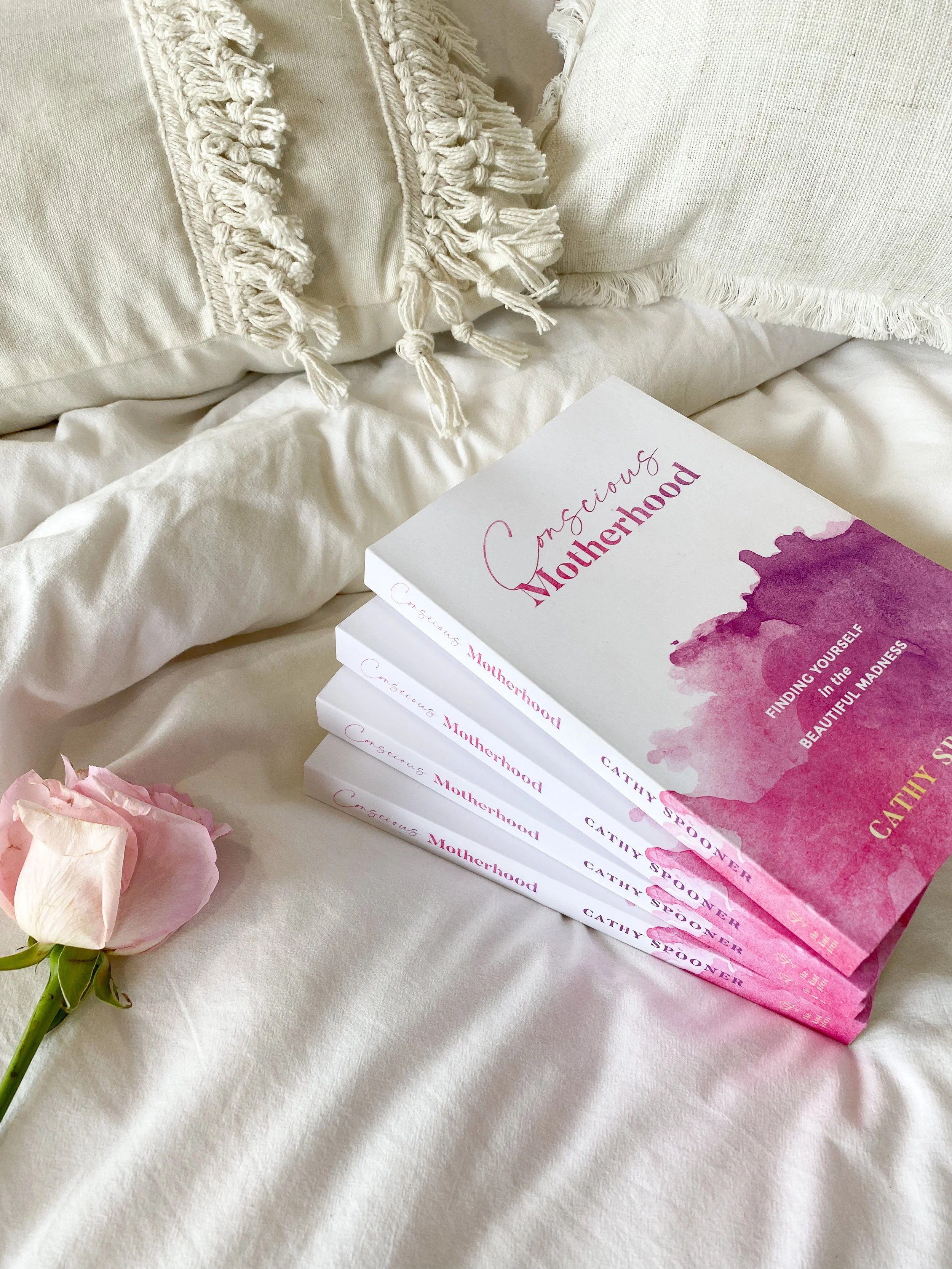 Stack of "Conscious Motherhood" books on a bed with a pink rose and white pillows.
