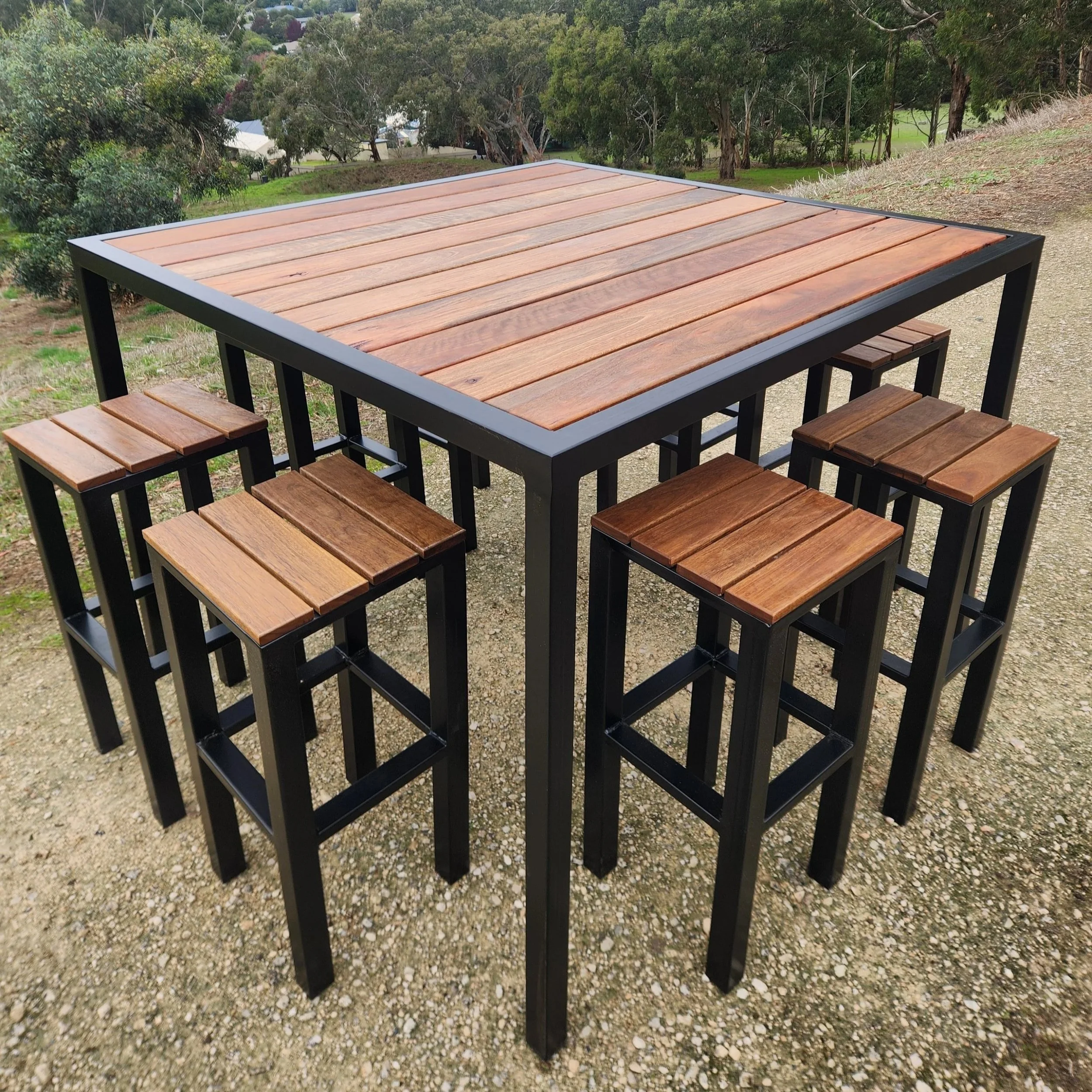Outdoor table with wooden slats and black metal frame, surrounded by matching benches, on a gravel surface with trees and houses in the background.