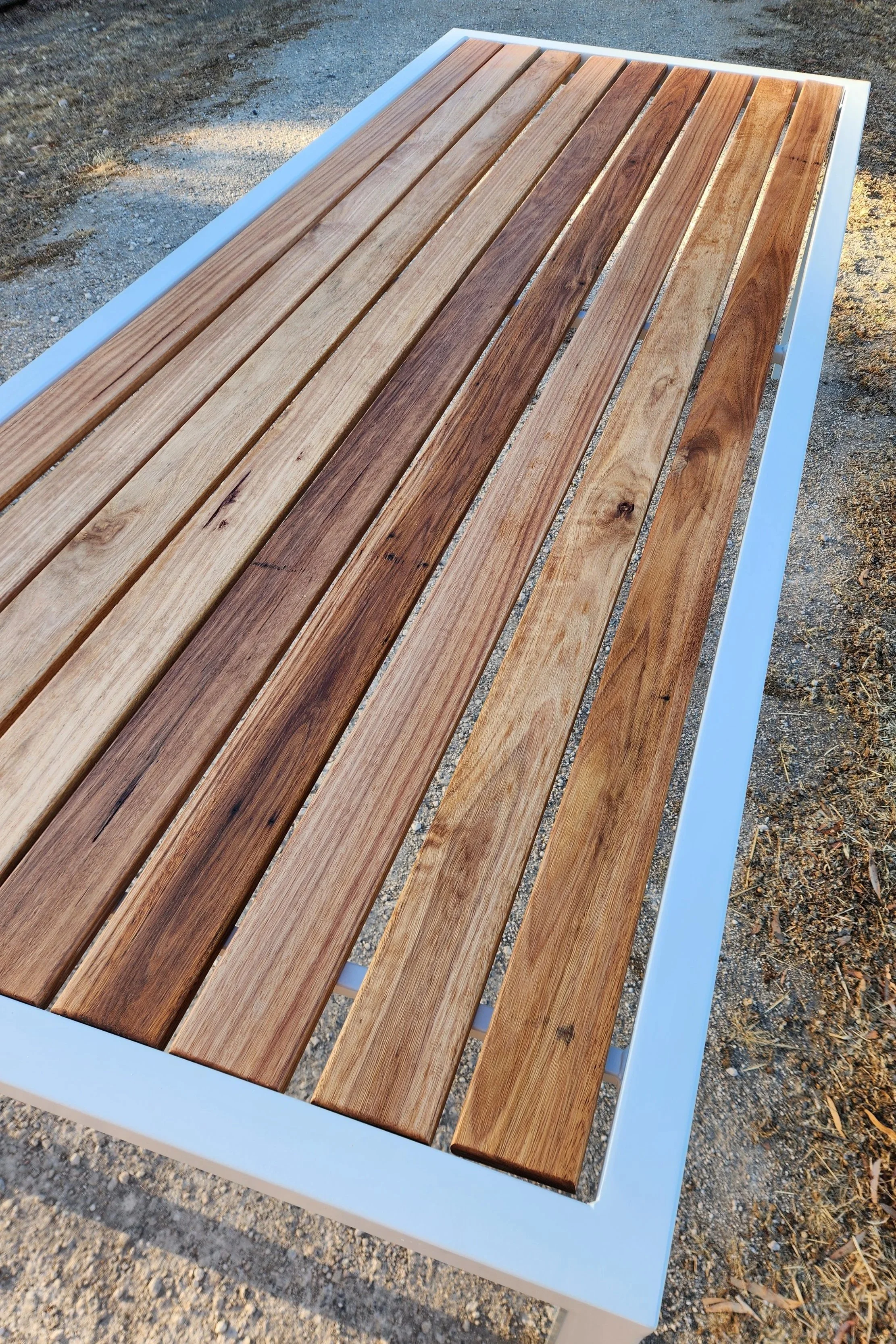 A rectangular outdoor bench with wooden slats on the seat and a white metal frame, placed on a gravel surface.