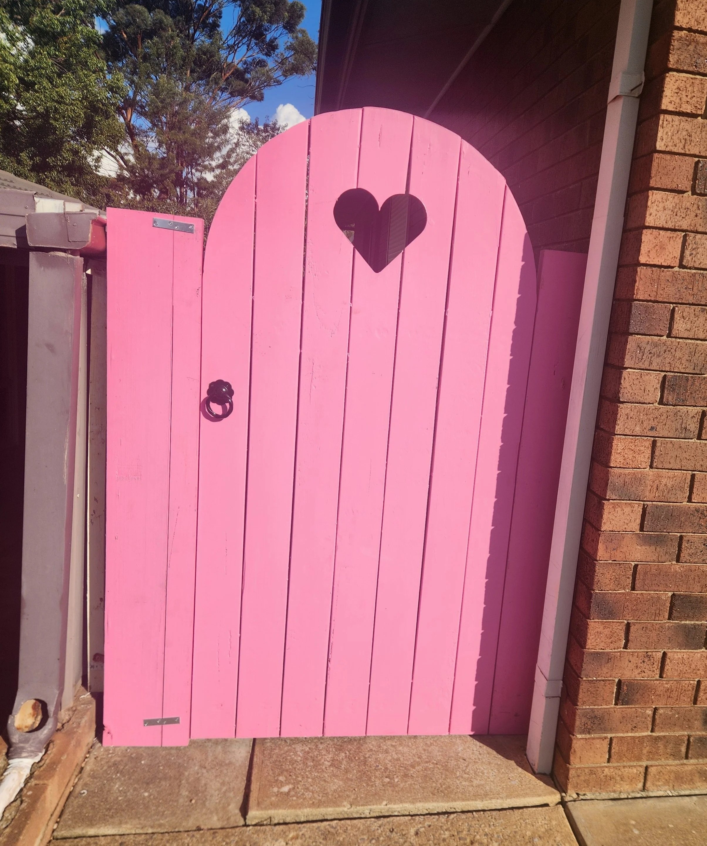 A pink wooden gate with a heart-shaped cutout, mounted on a brick wall, with a ring door knocker at the bottom, and a small black and white plaque at the lower left corner.