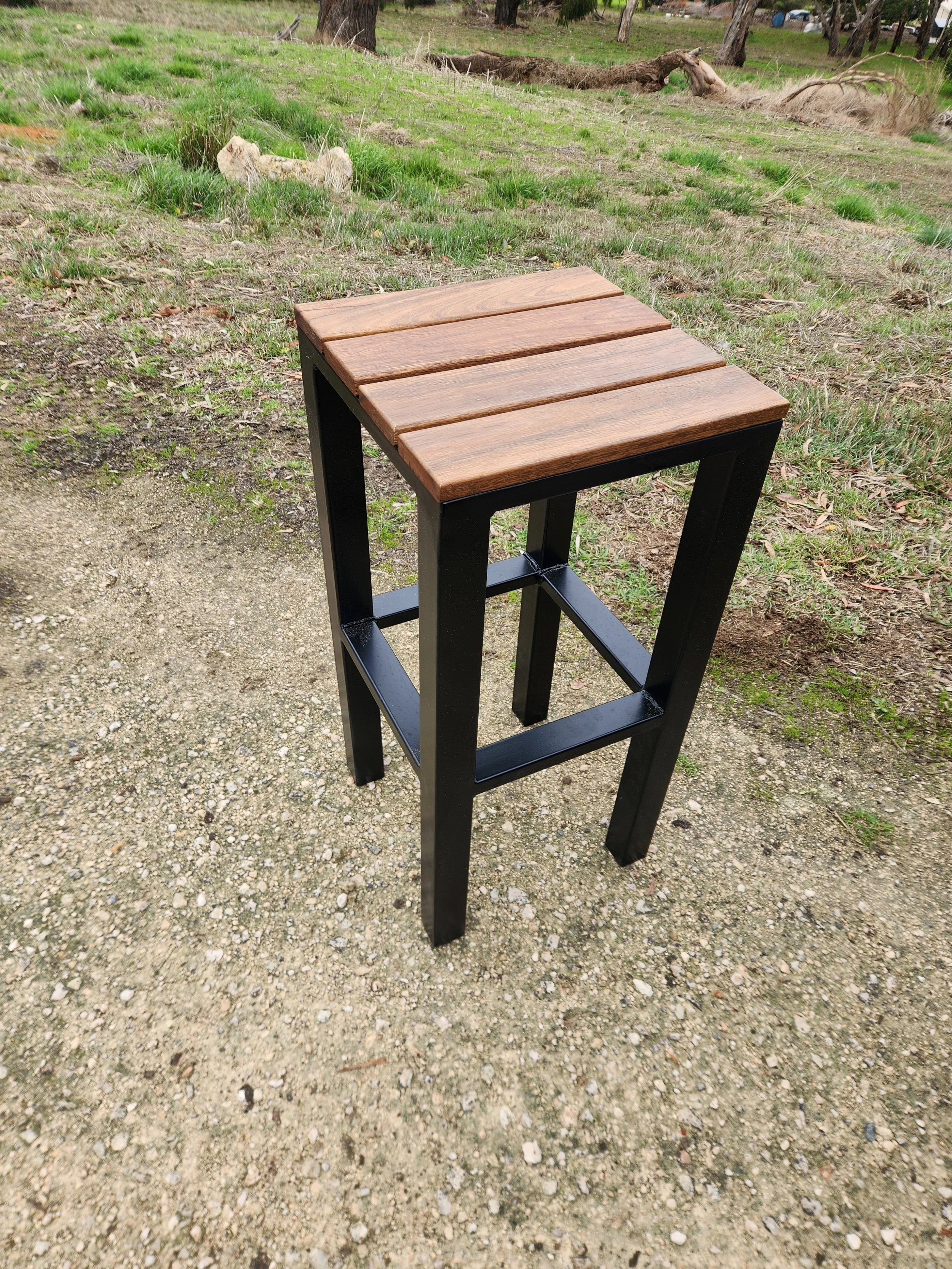 A small outdoor table with a wooden top and black metal frame, positioned on a dirt and gravel path in a grassy, wooded area.