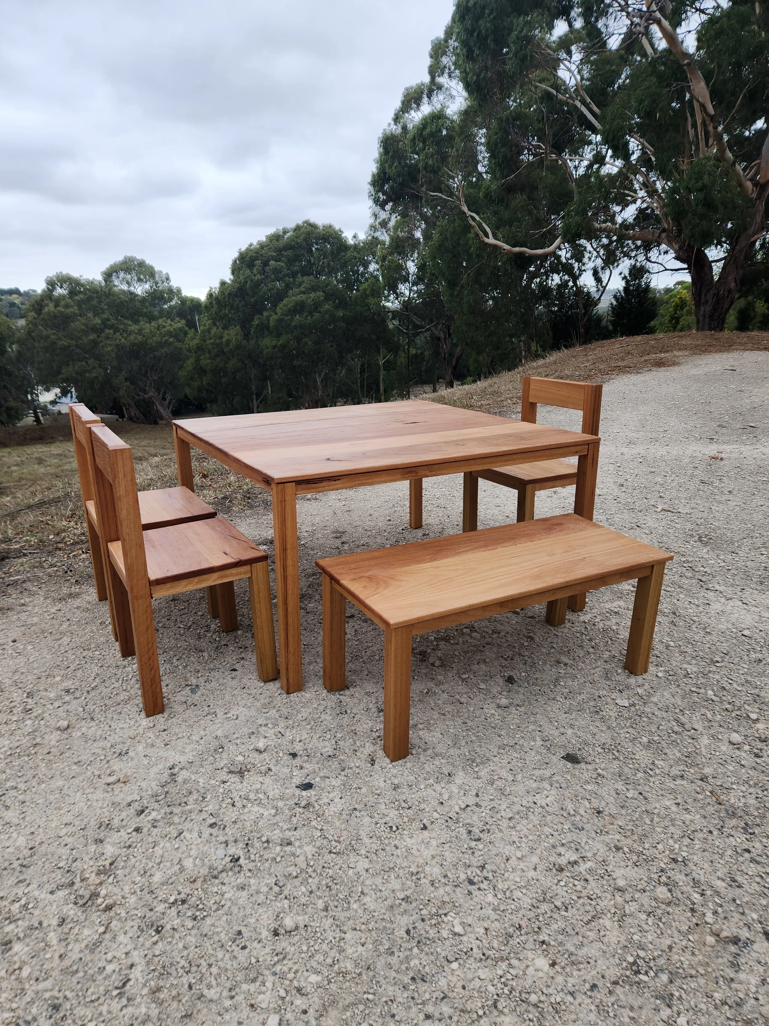 A wooden table with four matching wooden chairs is placed outdoors on a gravel surface. There are trees with green foliage and cloudy sky in the background.