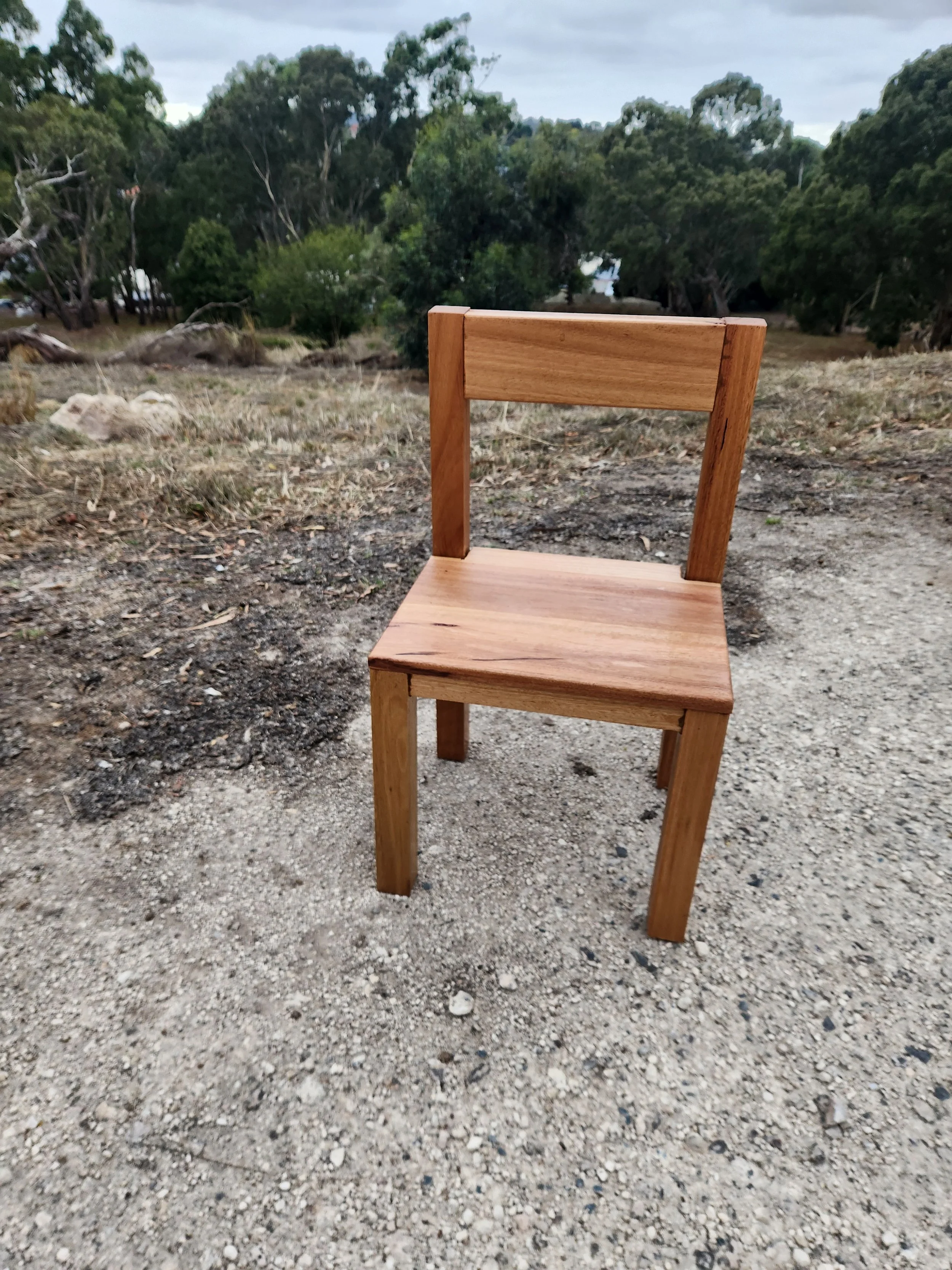A wooden chair with a backrest, placed on dirt ground outdoors with trees and a cloudy sky in the background.