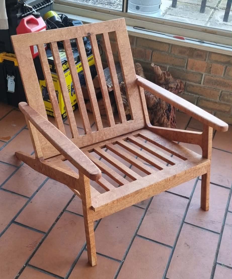Unfinished wooden outdoor bench with armrests placed on a tiled patio near a window, with a cat resting behind it and various tools and storage boxes in the background.