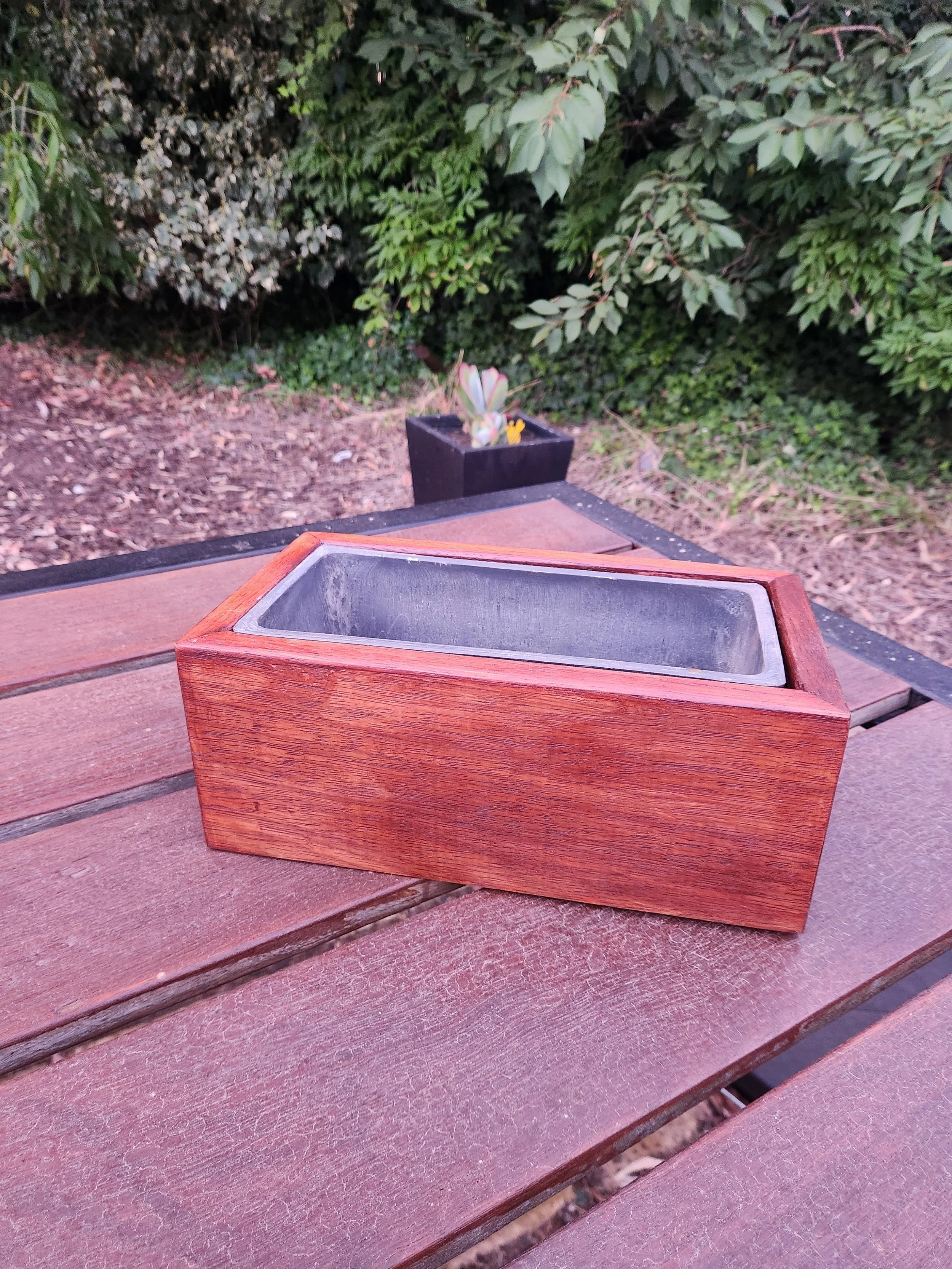 A wooden planter box with a metal liner is placed on a brown outdoor table, with a small black pot containing some plants in the background, and a lush green bush behind that.