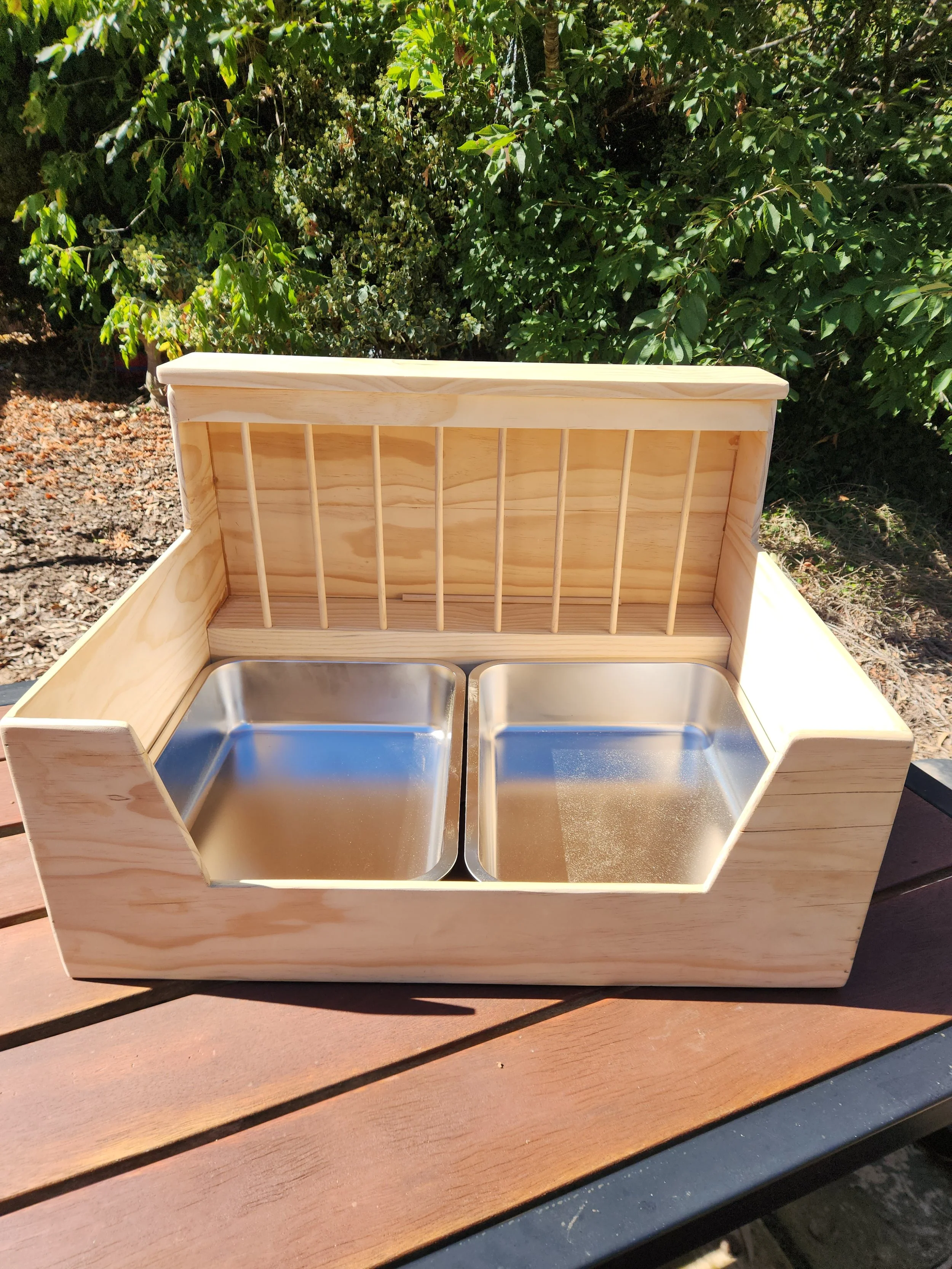Wooden kitchen sink cabinet with two stainless steel sink basins and a slatted wooden backsplash, outdoors on a wooden table with a background of green foliage.