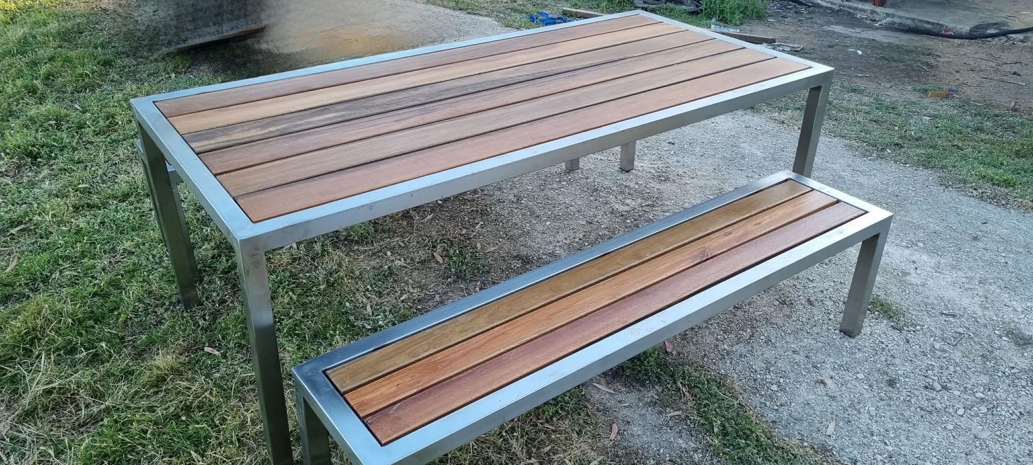 A modern outdoor bench with a metal frame and wooden slats, placed on a grassy and gravel surface.