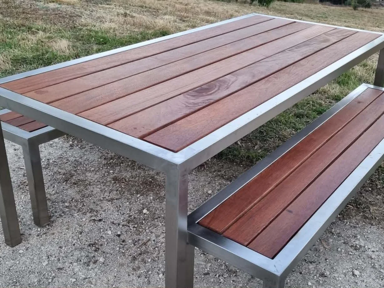 Outdoor metal table with wooden top and matching bench on a gravel surface, with grass and trees in background.
