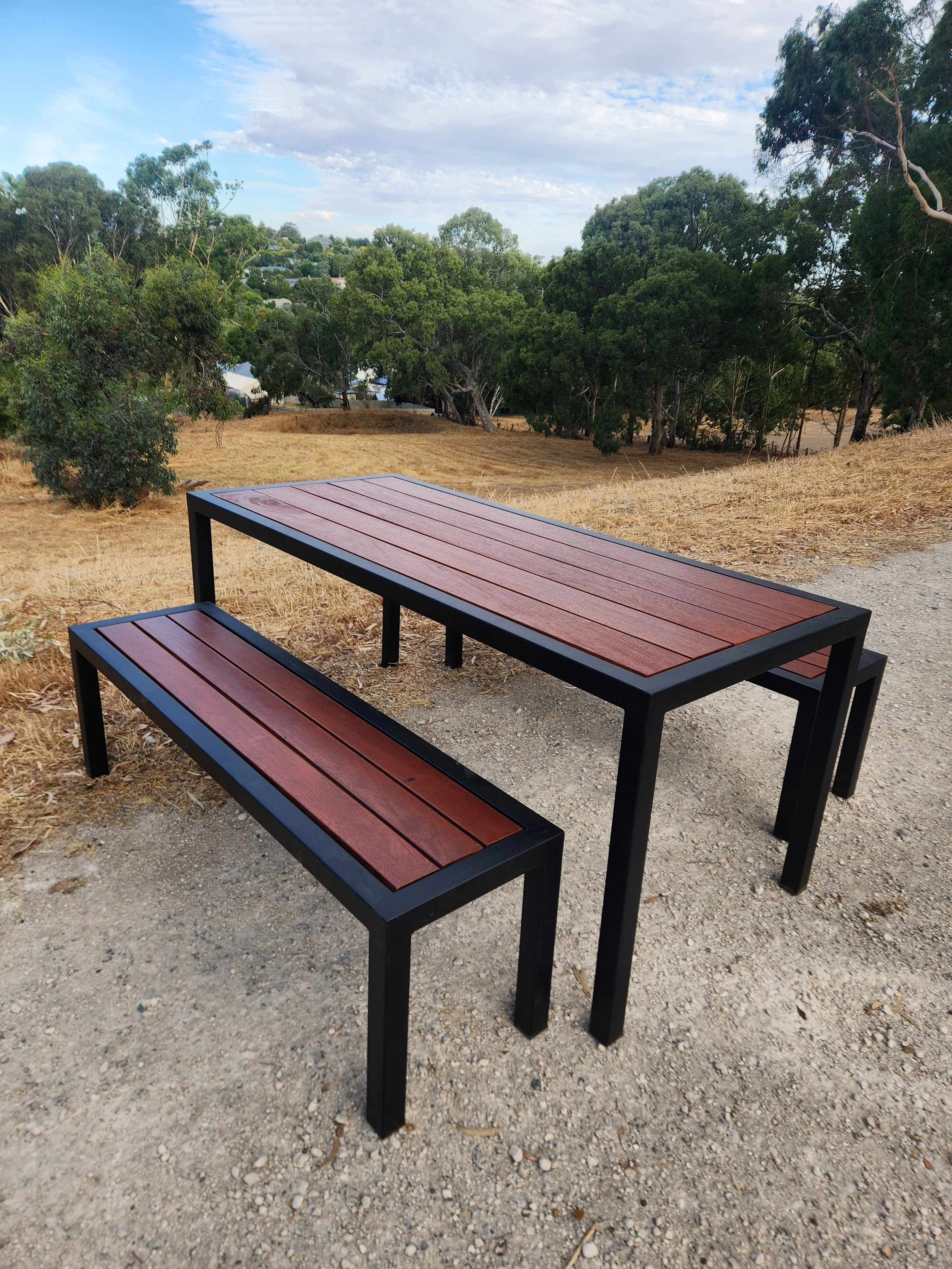 Outdoor scene with two wooden benches with black metal frames on a dirt path, trees and a cloudy sky in the background.