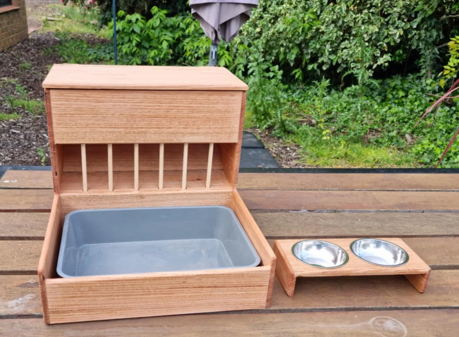 A wooden outdoor pet feeding station with a large box, a metal tray inside, and a smaller wooden stand holding two stainless steel bowls, set on a wooden picnic table with a garden background.