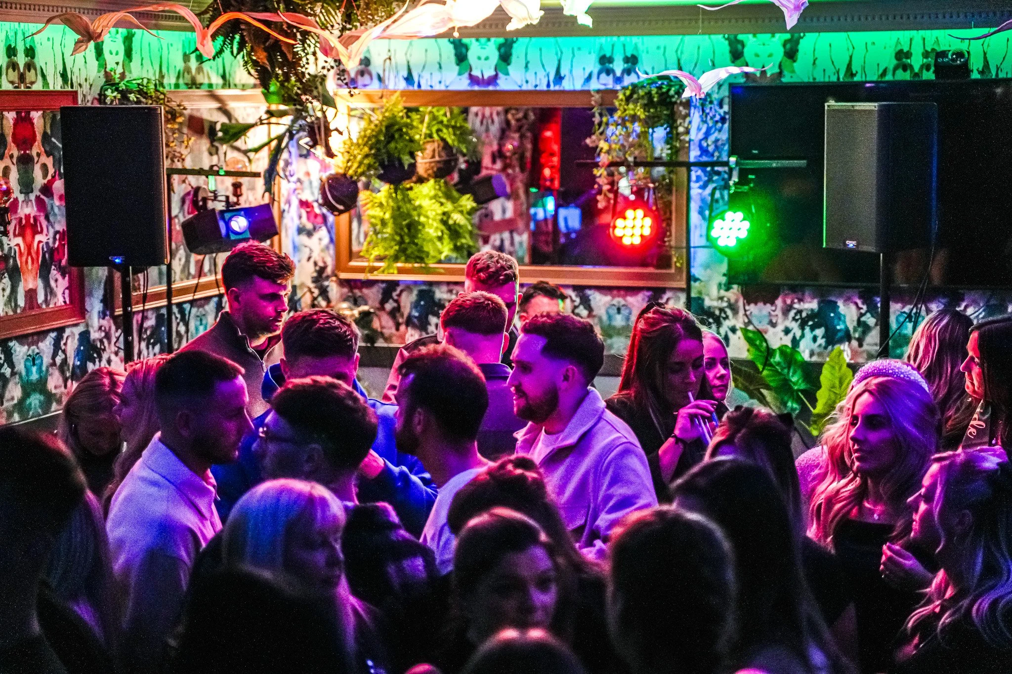 Crowd of young people dancing and socializing in a nightclub with colorful lighting, decorated walls, and hanging plants.