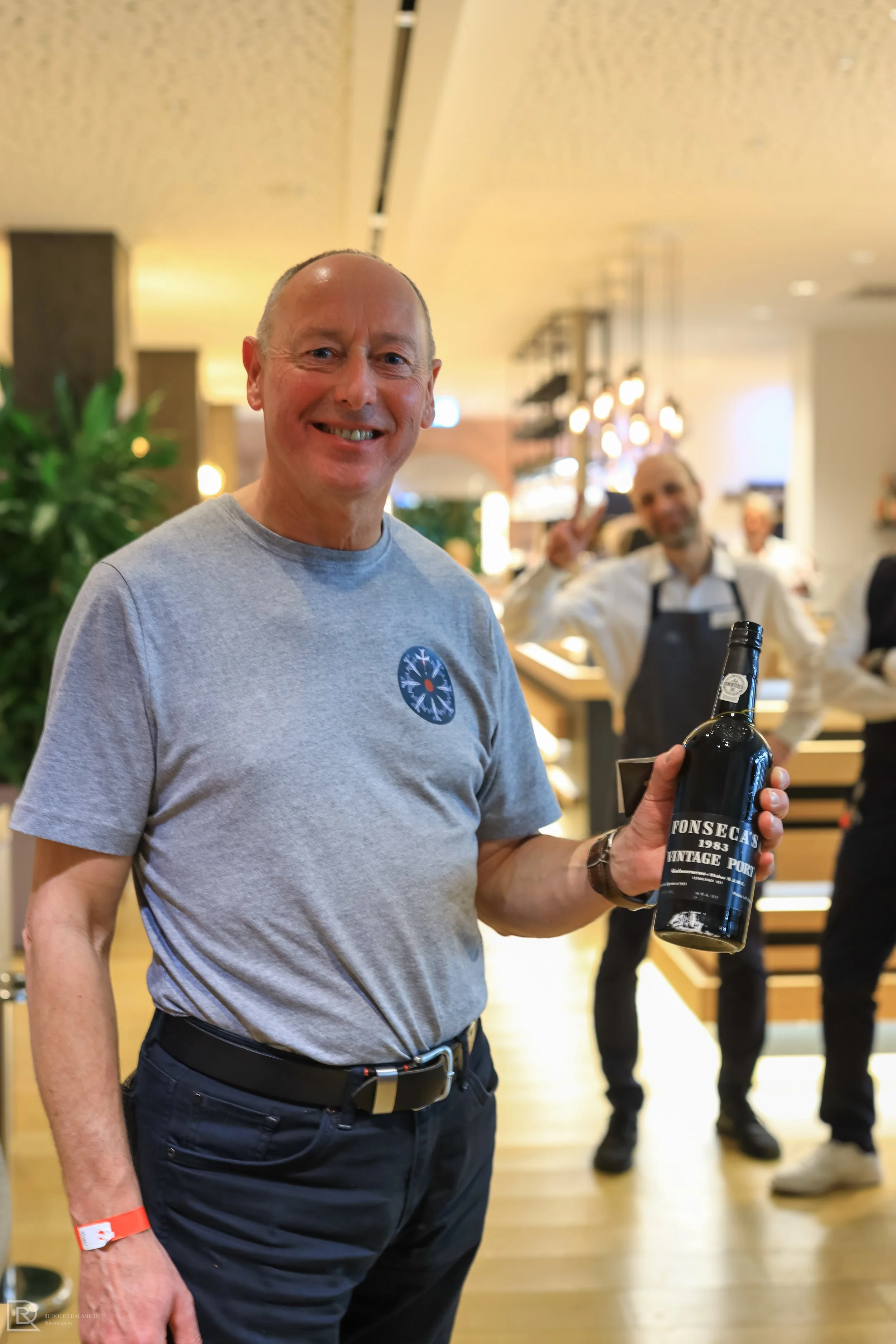 Man holding a bottle of Fonseca's Vintage Port 1983 in a modern indoor environment with people in the background.