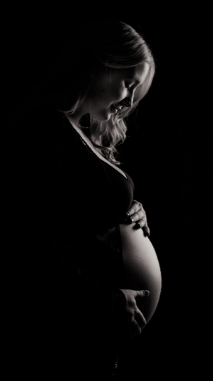 black and white image of a pregnant woman. side view. Her hands cradle her bare belly and she is gazing down at her growing bump with a content smile on her face.