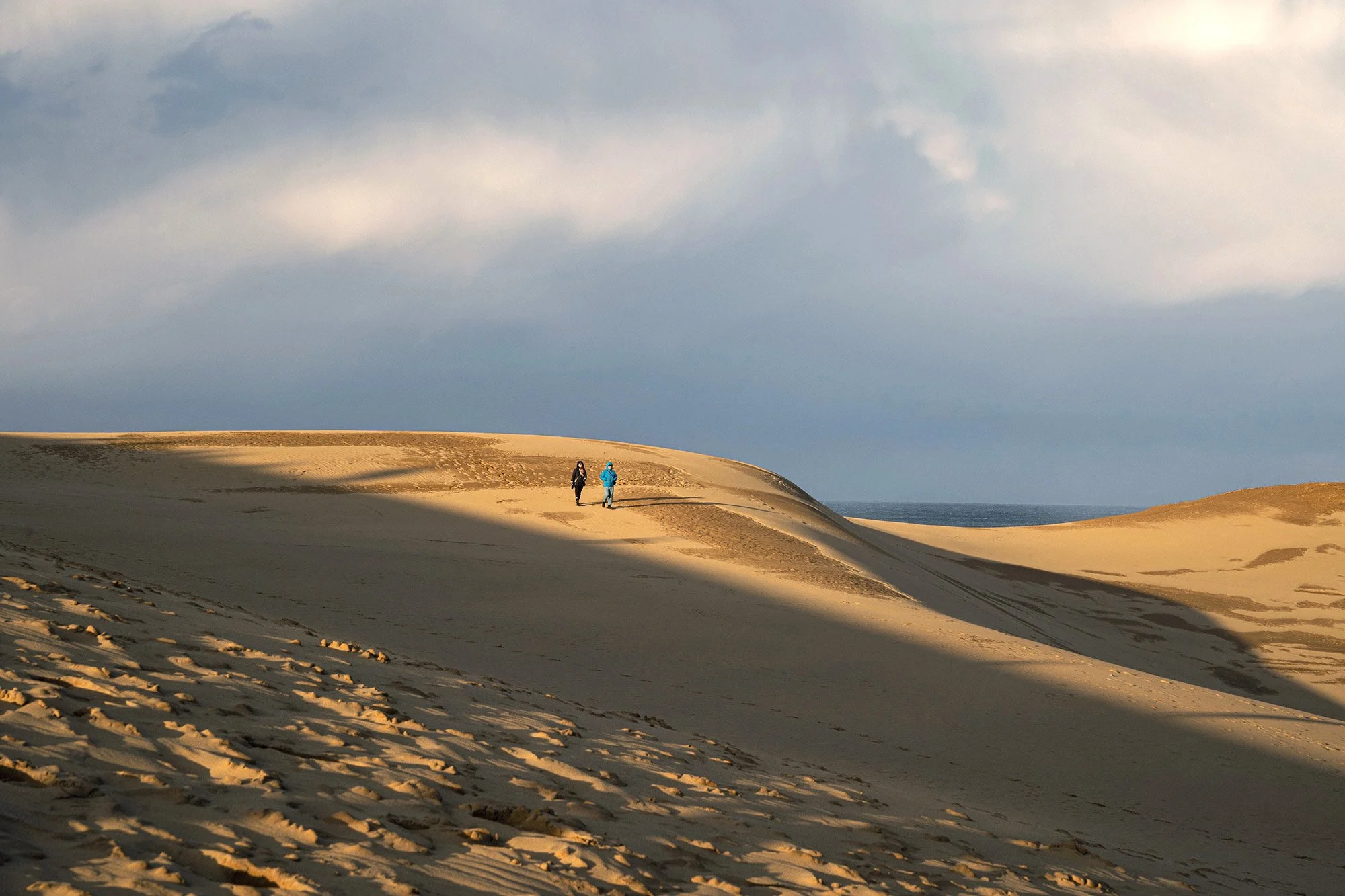 two people wearing jackets walking on sand dunes in the sunlight in tottori japan