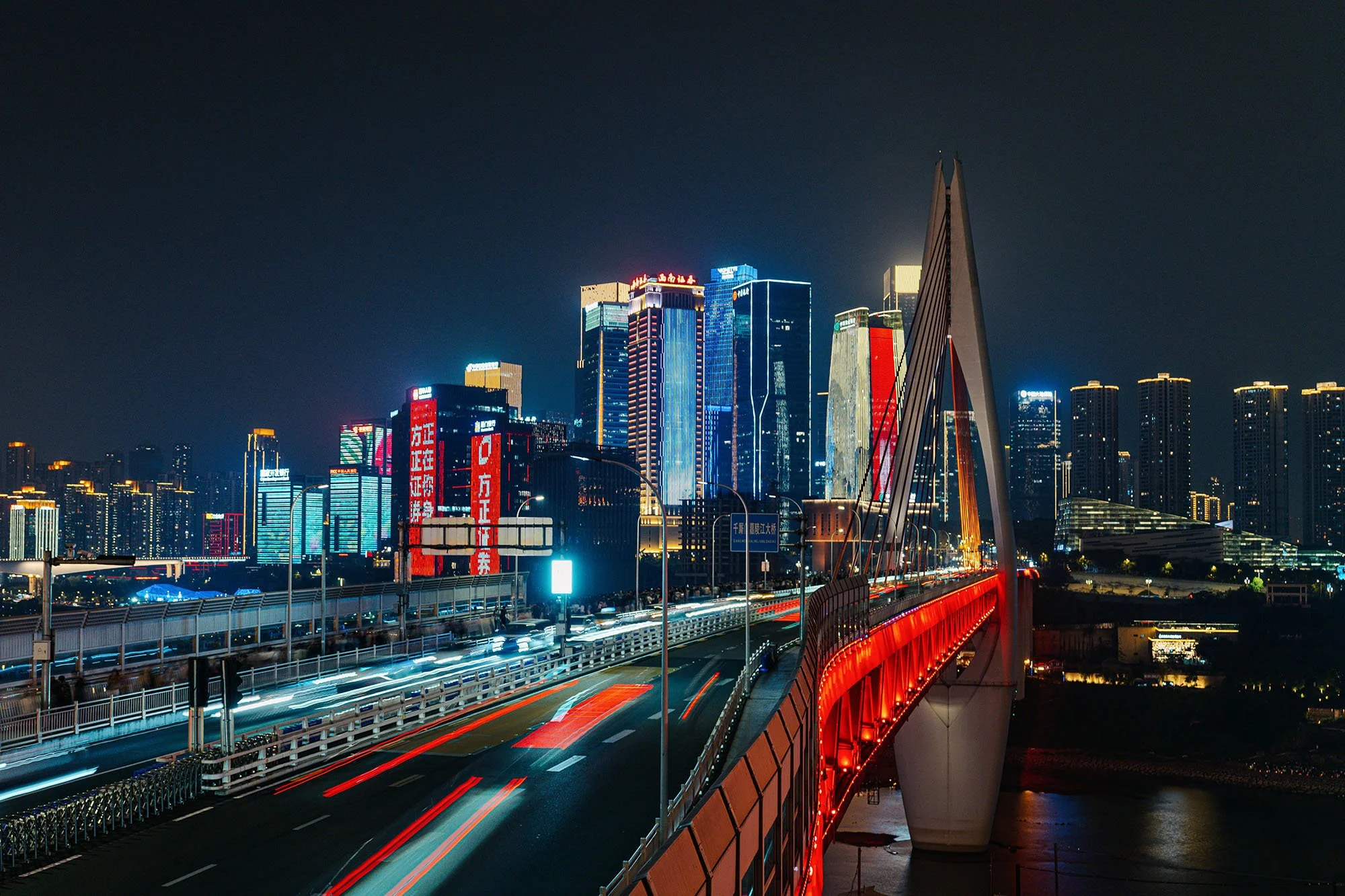 a bridge with red illuminations on its side crossing a river with car lights streaking on it and a skyline of skyscrapers illuminated in the background at night in chongqing china