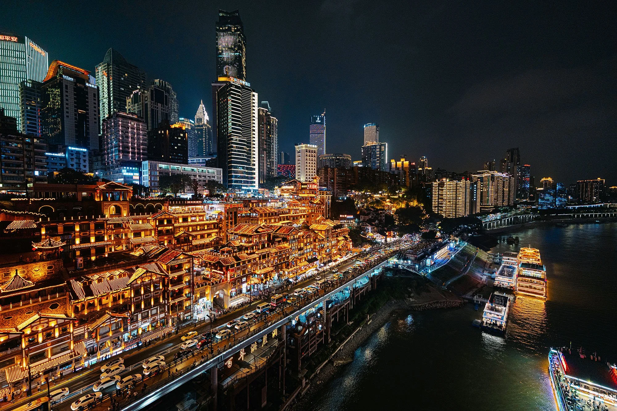 traditional chinese buildings illuminated in a golden light alongside a riverside at night with brightly lit skyscrapers behind it